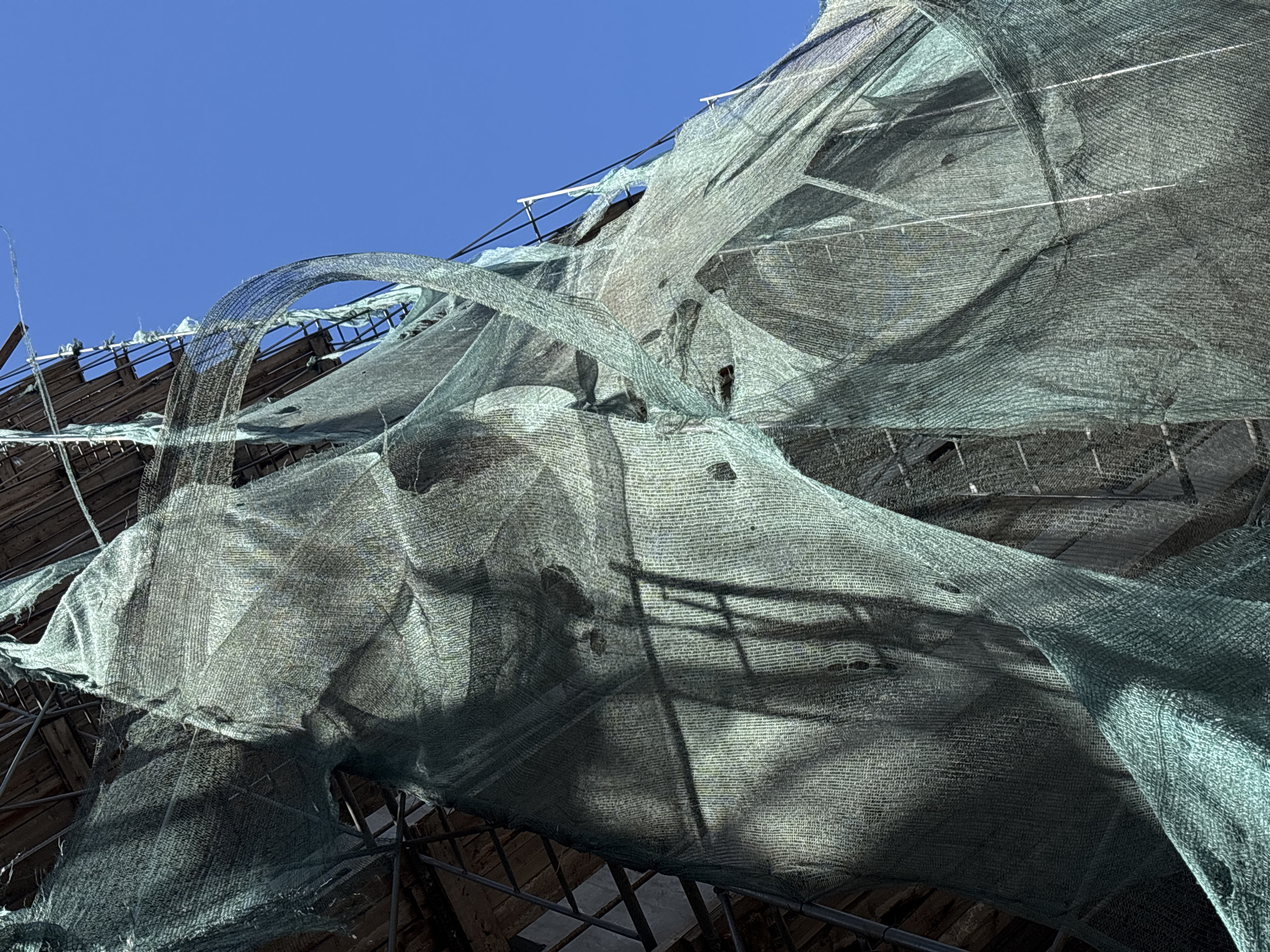 A low-angle shot depicts a building under repair, likely in the city of Unknown, Unknown. The building's skeletal wooden frame and metal scaffolding are partially obscured by a large, draped net of green netting. The netting is torn in places, allowing glimpses of the structure beneath. The clear, blue sky is visible in the upper left corner of the frame.