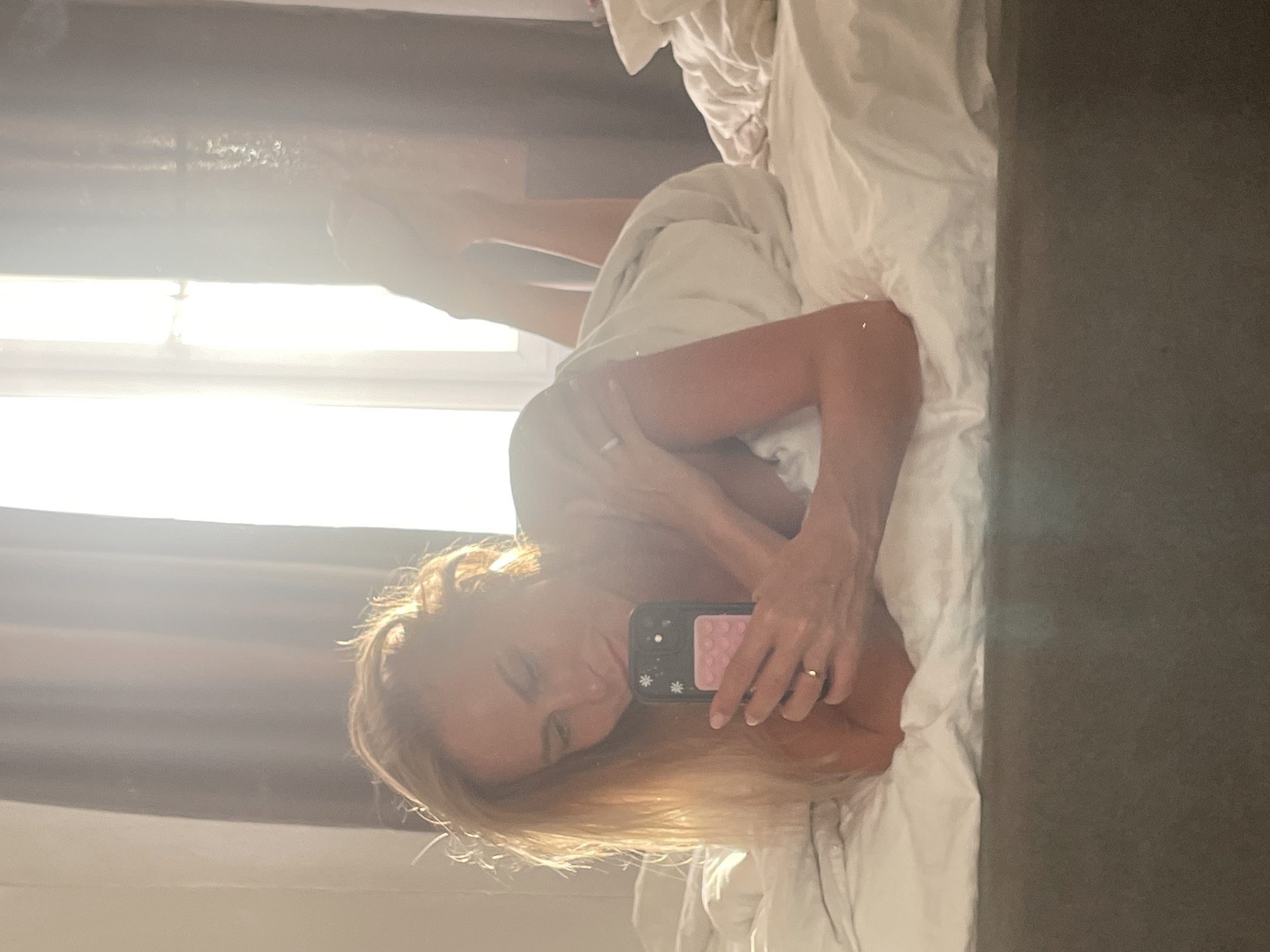 A blonde woman is lying on a bed, taking a selfie with her phone. She is wrapped in a white blanket, and her legs are propped up in the air. Sunlight streams in from a window behind her, creating a bright, airy atmosphere. The room appears to be a bedroom, with a large bed covered in white linens. The overall impression is one of relaxation and intimacy.