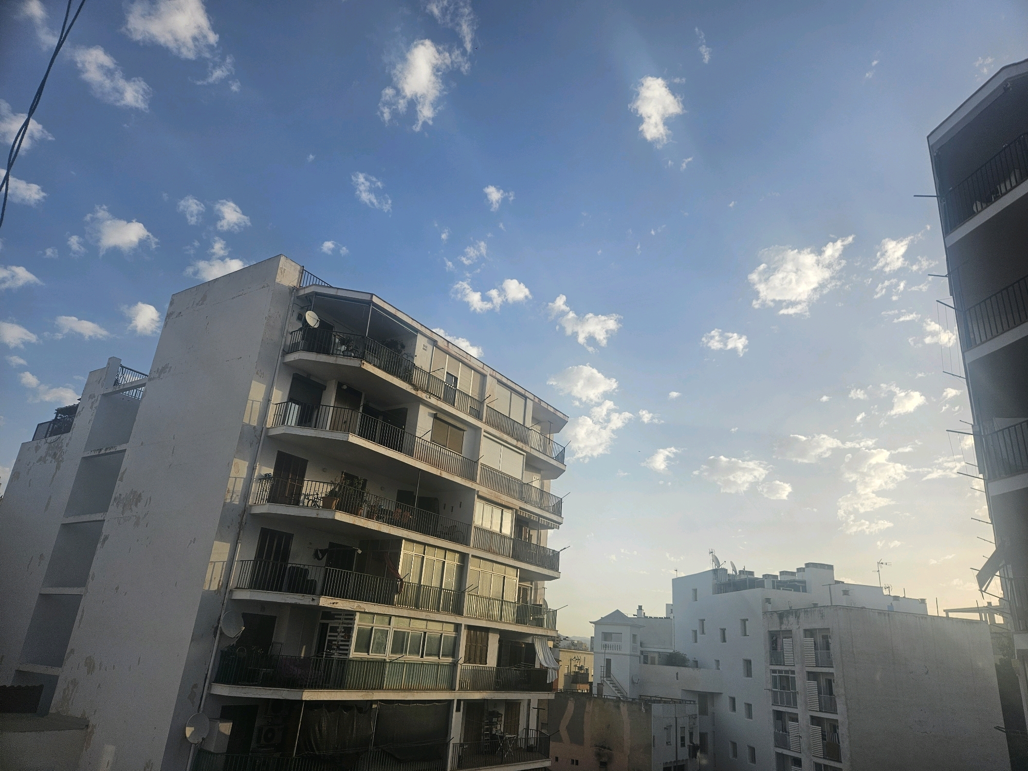 The image captures an urban scene in Palma de Mallorca, Spain, focusing on apartment buildings under a blue sky with scattered white clouds. The perspective is from a slightly elevated viewpoint, looking upwards and across at the facades of several multi-story residential buildings.
The most prominent building, in the foreground to the left, is a white, weathered apartment block with multiple balconies. The balconies are outlined by dark railings and feature various items such as satellite dishes, plants, bicycles, and clothes hanging to dry. The building's facade shows signs of age with peeling paint and visible structural elements. Sunlight casts shadows and highlights on the building's surfaces, suggesting it is either early morning or late afternoon.
To the right, another apartment building, darker in color, is partially visible, also with balconies and railings. Further in the background, a cluster of modern white buildings is seen, some with balconies and air conditioning units. One building stands out with a distinct tower-like structure at its top.
The sky is a vibrant blue, filled with soft, white cumulus clouds that are illuminated by sunlight. The overall impression is of a typical residential area in a coastal Mediterranean city, with a sense of everyday life unfolding. There are no people directly visible in the image, but the presence of personal belongings on balconies suggests the buildings are inhabited. The environment appears to be a densely populated urban area with buildings closely situated.
