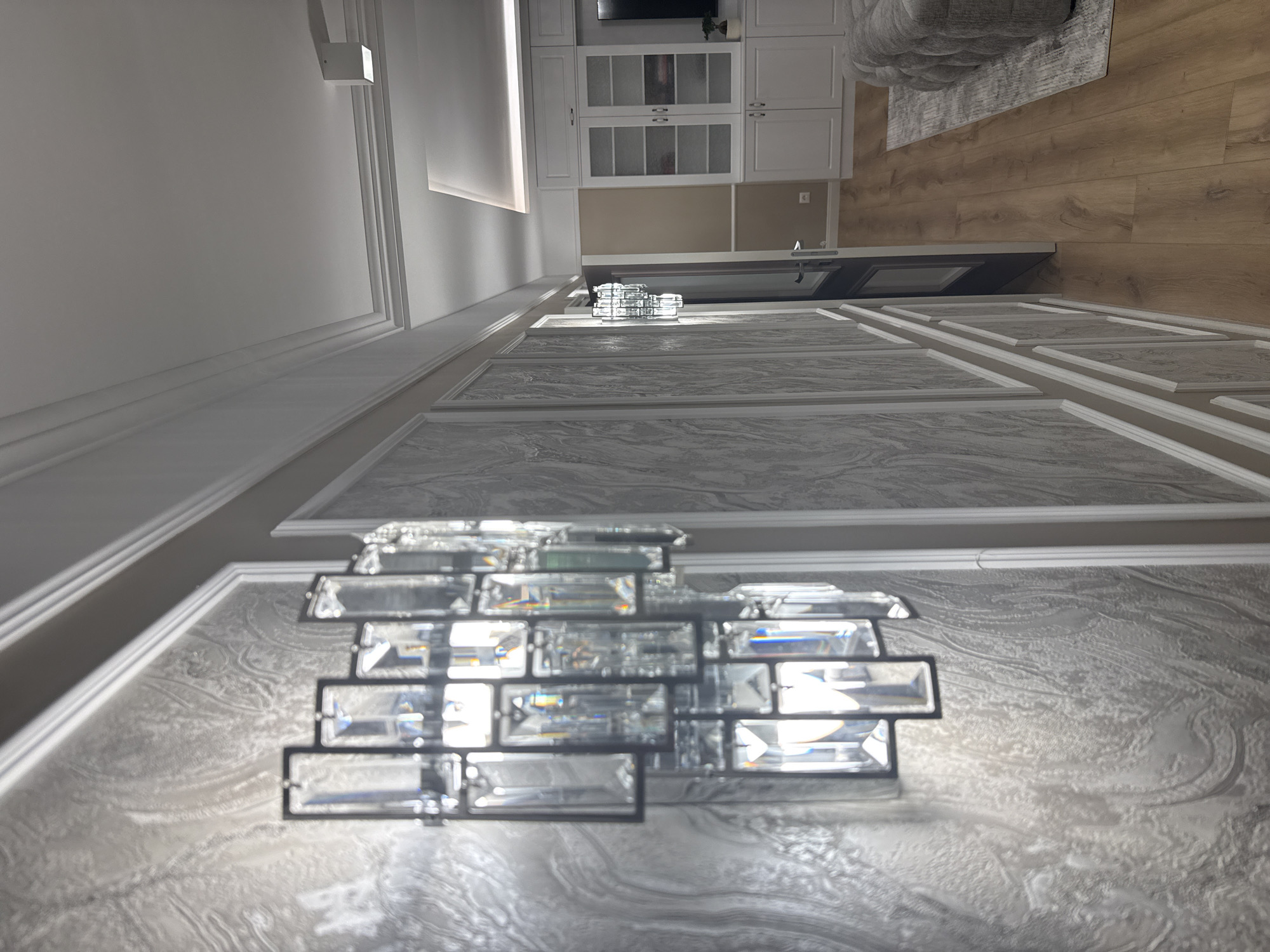 The image shows an interior space with a focus on the ceiling and lighting fixtures. The ceiling is decorated with framed panels featuring a marble-like pattern in shades of gray and white. Two crystal light fixtures are visible, one in the foreground and another smaller one in the background. The foreground light fixture is a modern design composed of rectangular crystal blocks arranged in a grid-like structure.
In the background, a kitchen area is partially visible with white cabinetry and a dark countertop. Part of a gray sofa and a rug are also seen. The floor is light wood.
Given the architectural details and the overall aesthetic, the image likely depicts a modern, upscale interior. Specific location within Sofia, Bulgaria cannot be determined from the image alone.