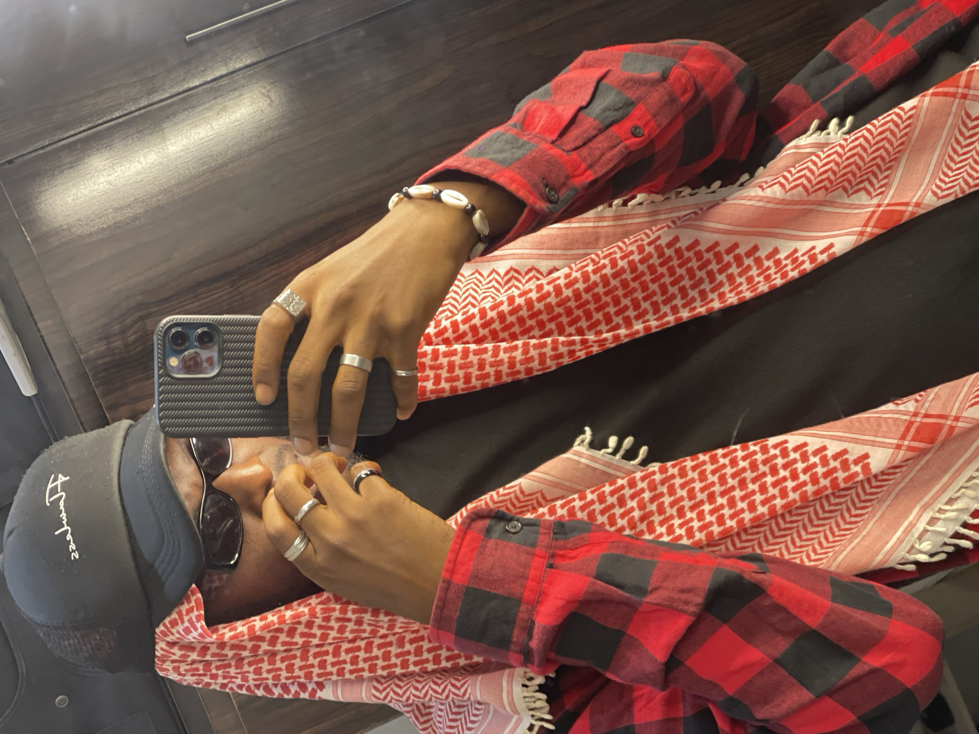 A person is taking a selfie in a mirror. They are wearing a black trucker hat with white lettering, sunglasses, a red and black checkered shirt, and a white and red keffiyeh. They have several rings on their fingers and a bracelet made of shells and beads on their wrist. The background appears to be a wooden closet or door. The lighting suggests an indoor setting, possibly a bedroom or dressing area. The image is taken at eye level, with the person holding a smartphone to capture the reflection. The overall scene is casual and personal.