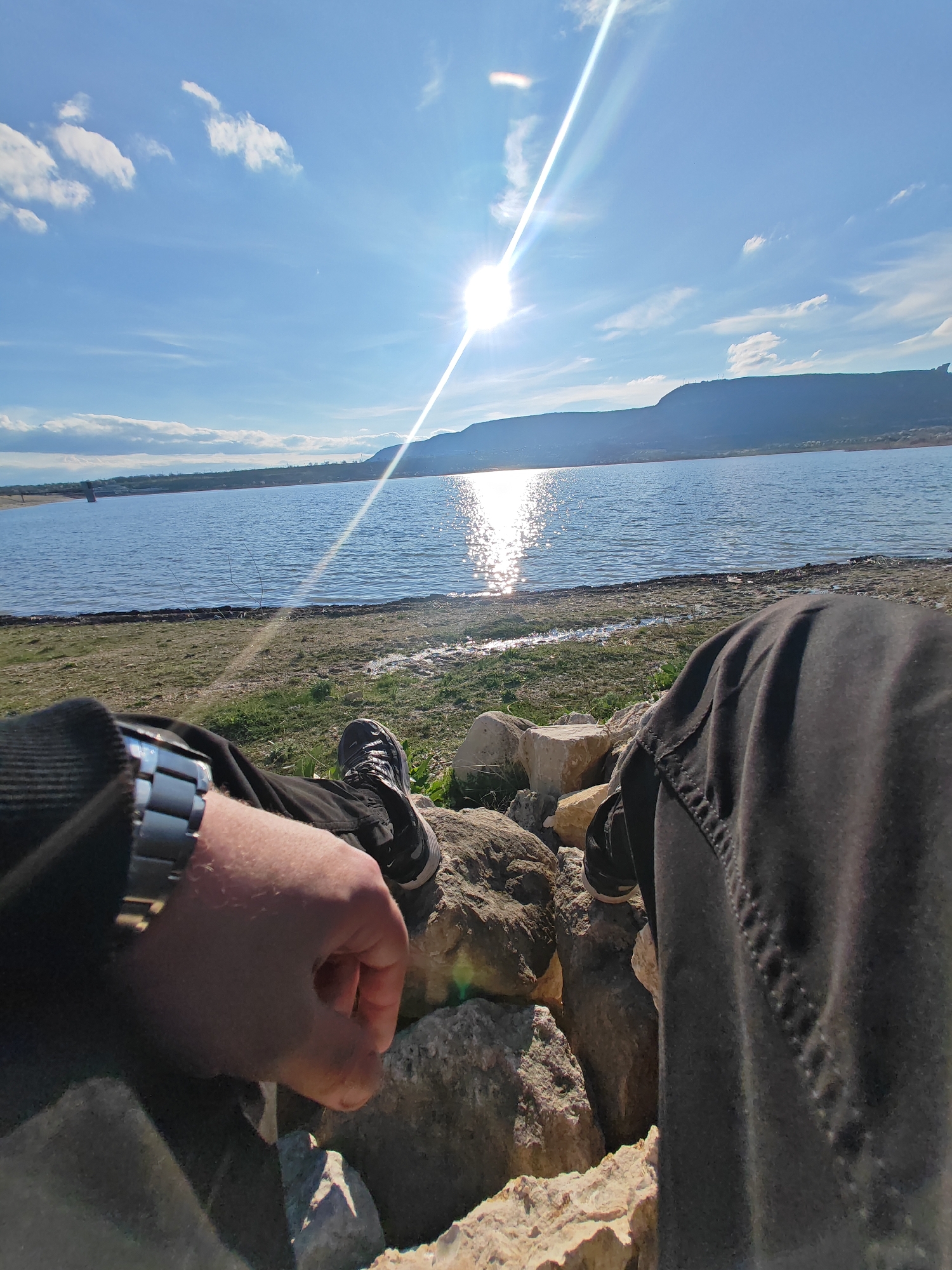 A first-person, low-angle shot shows a person sitting on rocks near a body of water under a bright, sunny sky. The person is wearing black pants and black sneakers. Their left arm is visible, adorned with a silver wristwatch. The water reflects sunlight, creating a bright glare. In the background, a distant land mass is visible across the water. The sky is blue with scattered clouds. The rocks are light-colored and vary in size. Some green vegetation is present on the shore. The location is near the water reservoir of the city of Shumen, Bulgaria.