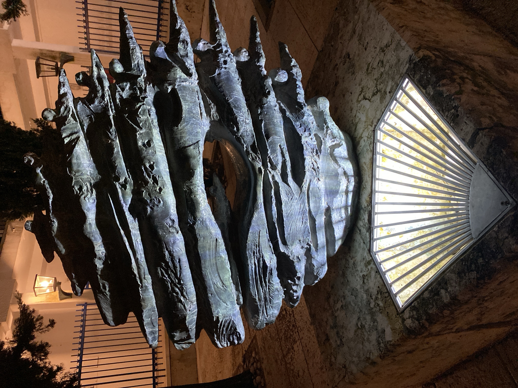 The image depicts a nighttime scene in Budva, Montenegro, featuring the abstract sculpture "Seaside Melody" by Kosta Bogdanović, located near the Old Town promenade. The central subject is a large, multi-layered sculpture crafted from dark metal, likely bronze or oxidized copper, presenting an undulating, wave-like, or stratified geological appearance. Approximately ten distinct horizontal layers are visible, with several stylized human figures or heads integrated into and emerging from the upper sections of the sculpture. The sculpture rests on a multi-tiered, light-colored concrete or stone pedestal.
Directly in front and below the sculpture, embedded within the pedestal, is a fan-shaped light fixture composed of white vertical bars over a yellowish-translucent panel. This fixture emits a bright, yellowish-white light upwards, illuminating the underside and intricate textures of the sculpture with cool blueish-white reflections. Further ambient light is provided by two wall-mounted lanterns with frosted glass panels, casting a warm orange glow, attached to a light-colored building positioned in the background. A dark metal fence with vertical bars runs horizontally behind the sculpture, separating it from the building. On the left side of the frame, a dark tree branch with sparse foliage is visible against the background. The ground surrounding the pedestal appears to be paved with light-colored, possibly textured, material. No individuals are present in the scene.