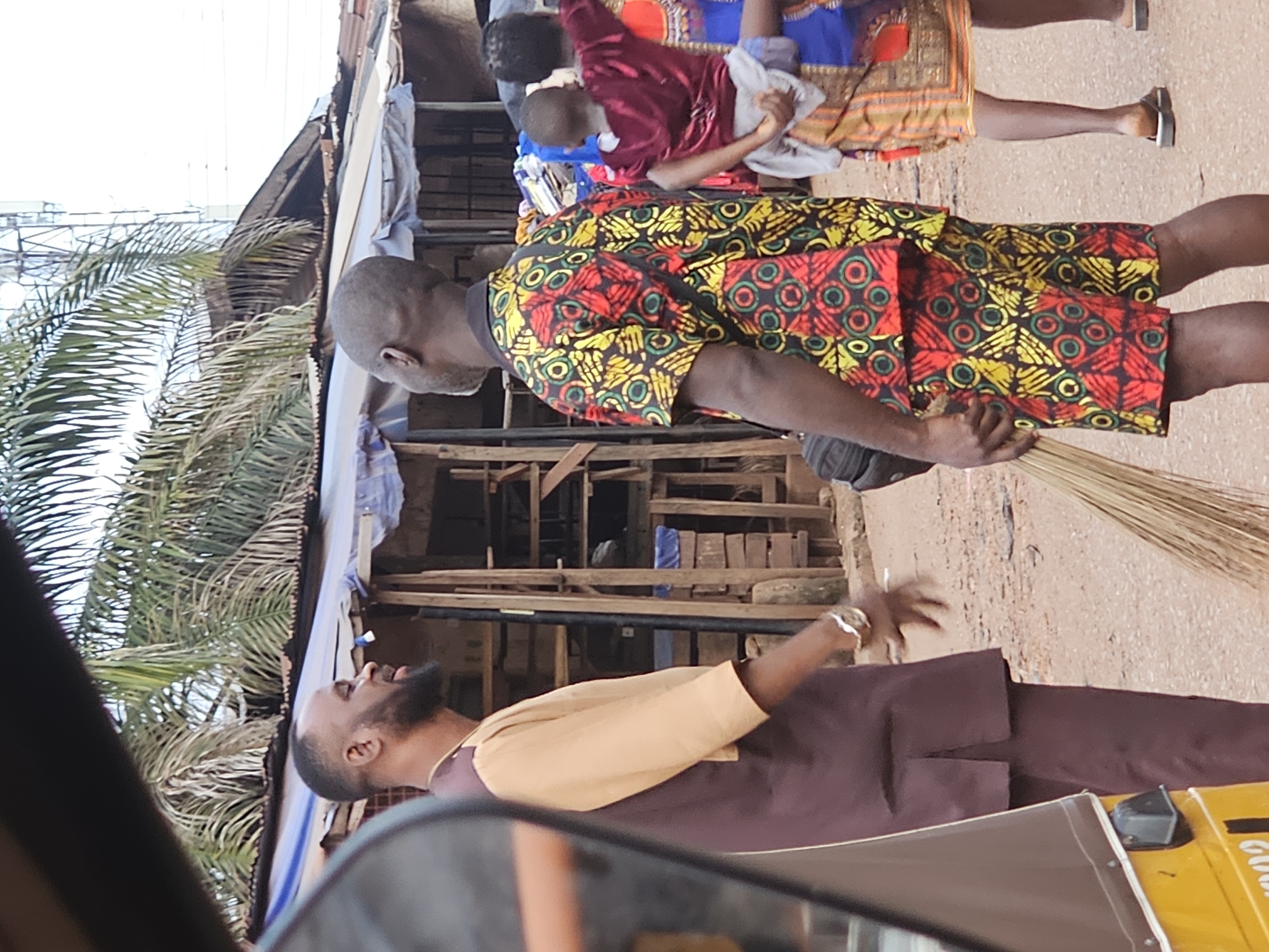 In an outdoor, dirt-road market scene, two men and a woman carrying a child are visible. The man on the left, with a beard and wearing a two-toned brown and tan tunic, appears to be speaking, with his mouth open and left hand raised, wearing a watch on his left wrist. To his right, a man wearing a brightly patterned short-sleeved shirt and shorts in red, yellow, and green, holding a broom, is facing away from the camera. Behind them, a woman with braided hair, also facing away, carries a child on her back, the child wearing a dark red shirt. The woman is wearing a patterned skirt in orange and blue. The background shows market stalls with corrugated metal roofs and wooden structures, with palm trees overhead and a cell tower visible in the distance. A yellow auto-rickshaw is partially visible on the left side of the frame.