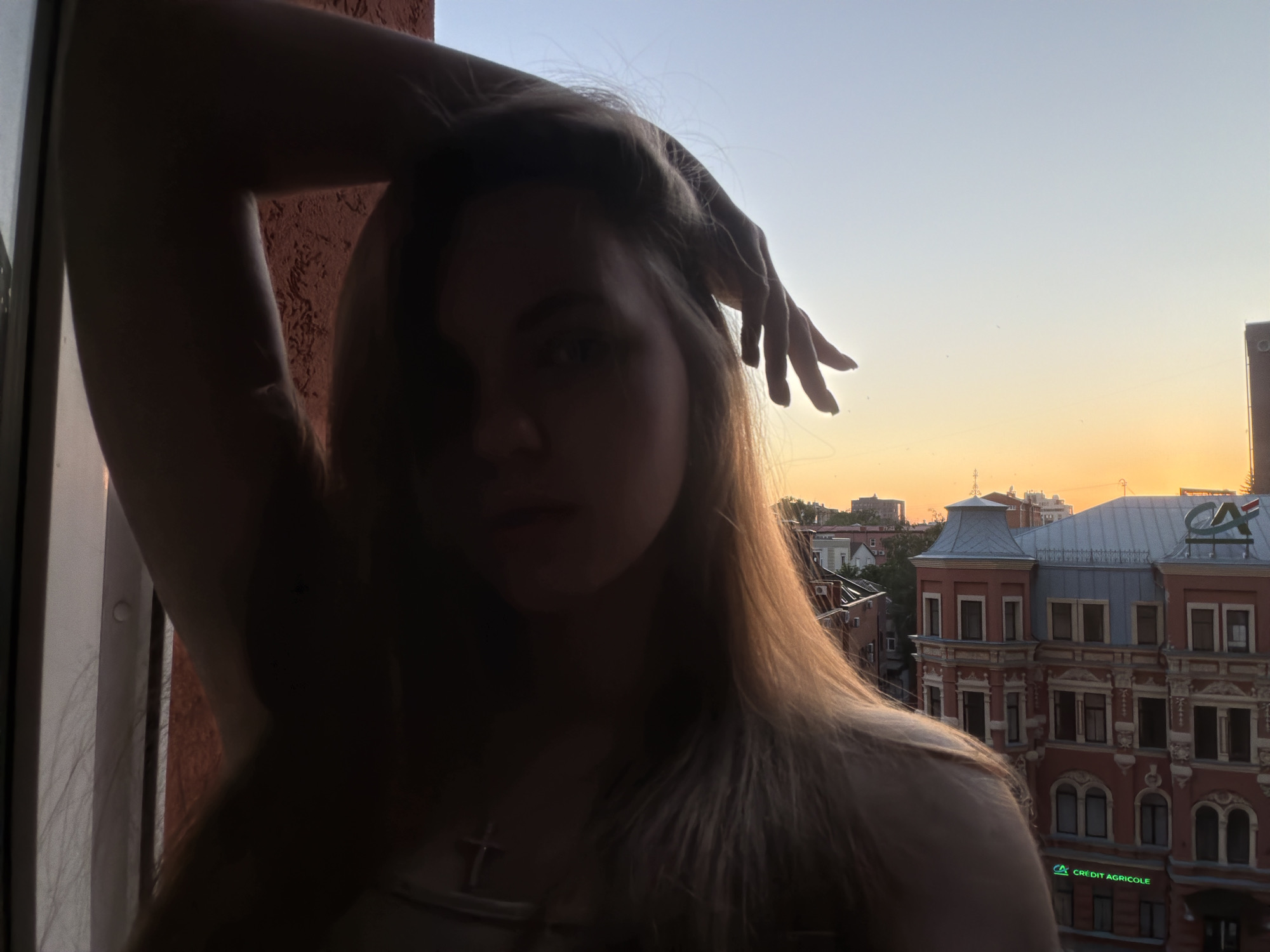 In an outdoor, urban setting during sunset, a young woman is positioned in the foreground, silhouetted against the orange and blue sky. Her left arm is raised above her head, and her hand rests on her hair. She is wearing a cross necklace. Behind her, an ornate red building with grey accents and numerous windows is visible. A green logo, "CREDIT AGRICOLE," is present on the building. The sky transitions from a clear blue at the top to a gradient of orange and yellow near the horizon, indicating the setting sun. The overall scene is one of a person enjoying a view from a window in a city.