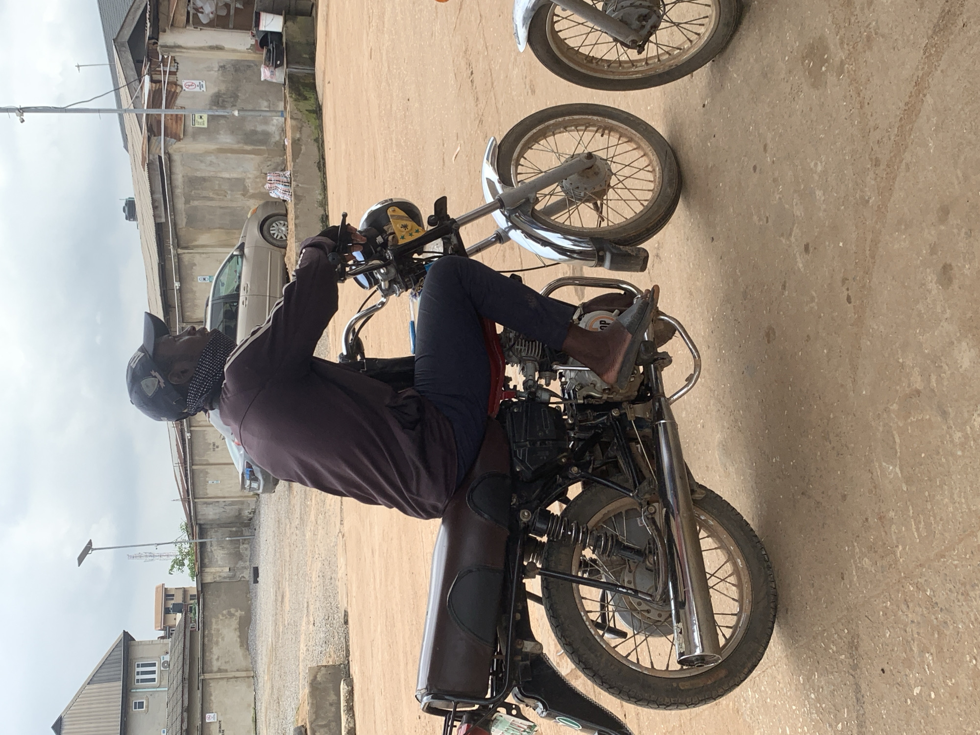 The image depicts a man riding a motorcycle on a dirt road in Shagamu, Nigeria. The man is wearing a dark long-sleeved shirt, dark pants, a dark baseball cap, and a patterned face mask. He is wearing sandals. The motorcycle is dark brown and appears to be a standard model. In the background, there are buildings with corrugated metal roofs, a beige car, and a fence. The sky is overcast. The scene appears to be a typical street view in a Nigerian city.