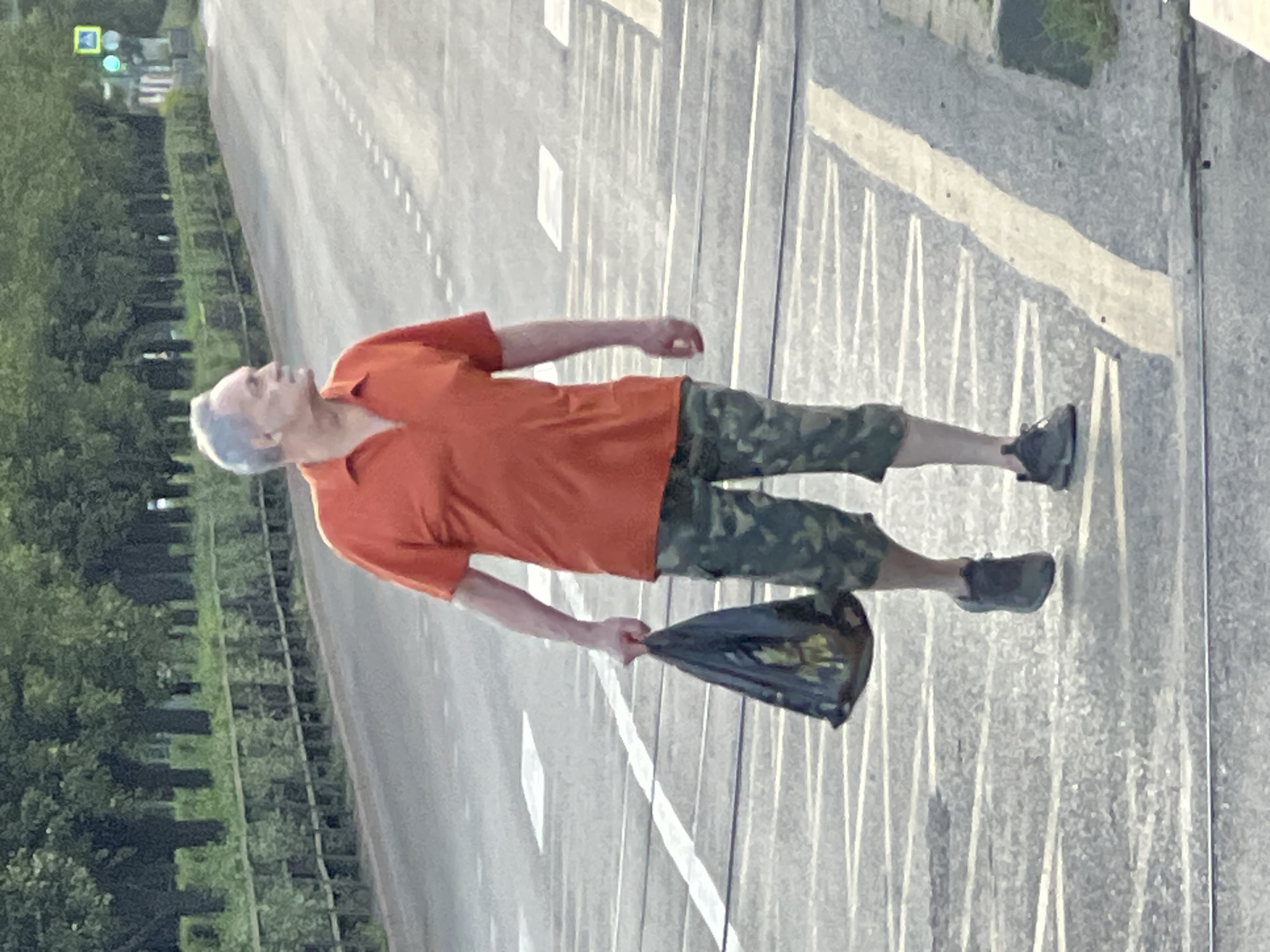 An elderly man with graying hair is walking across a crosswalk, carrying a black plastic bag in his left hand. He is wearing an orange polo shirt and camouflage shorts. The man is looking to his right. The crosswalk is on a paved road with white dashed lines indicating lanes. In the background, there are trees and a traffic light. The scene appears to be in an urban environment in Krasnodar, Russia.