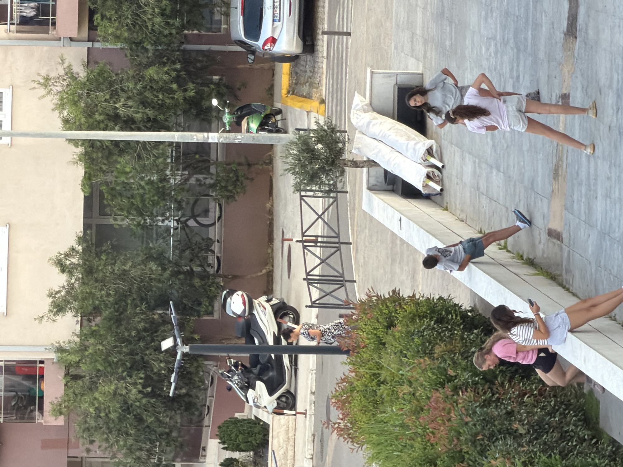 This media file shows an outdoor scene with several people and objects. The setting appears to be a street or walkway adjacent to buildings and lush greenery, possibly in Budva, Montenegro.
In the foreground, a white and black scooter is parked. Further up the walkway, a person in a black and white patterned dress is bent over, seemingly interacting with the scooter or something near it.
Further in the background, a silver car is parked. There are also two people, likely young women, walking or standing on a raised concrete structure. One is wearing a light grey long-sleeved top and shorts, while the other wears a white and pink striped top and shorts. Another person, wearing a grey t-shirt and blue shorts, is seen in a push-up position on the same elevated structure.
The scene is well-lit, suggesting it might be daytime, and the weather appears pleasant. The presence of trees and foliage indicates an outdoor environment. No specific emotions are overtly displayed by the individuals, but their casual attire suggests a relaxed atmosphere. There is some text visible on the rear of the silver car, a license plate with "BD 8E 568". The overall impression is a snapshot of everyday life in an urban or semi-urban setting.