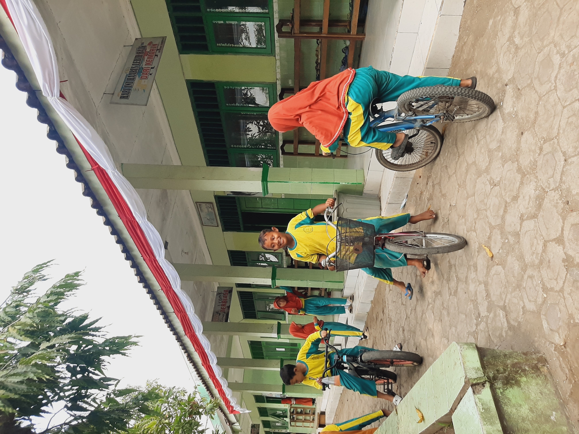 In front of a light green, one-story building with a red and white draped awning, several children wearing yellow and teal school uniforms are on bicycles on a light brown, hexagonal-patterned paved area. Some of the children are wearing headscarves. In the background, there are green-framed windows, wooden shelves, and a sign with Indonesian text. A leafy tree is visible in the upper left of the image. The scene appears to be a school in Sumberjo, Indonesia.