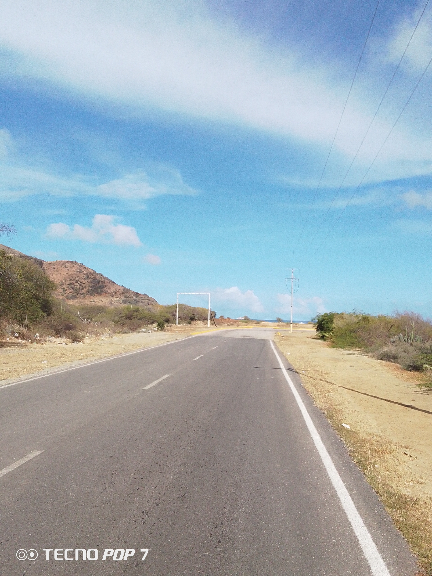 A desolate asphalt road stretches ahead under a partly cloudy blue sky in Pedro González, Venezuela. The road is marked with white lines and bordered by arid vegetation and scrubland. A low, brown hill rises in the distance to the left, while an electrical power pole stands along the right edge of the road. A rectangular white archway is set on the road ahead.