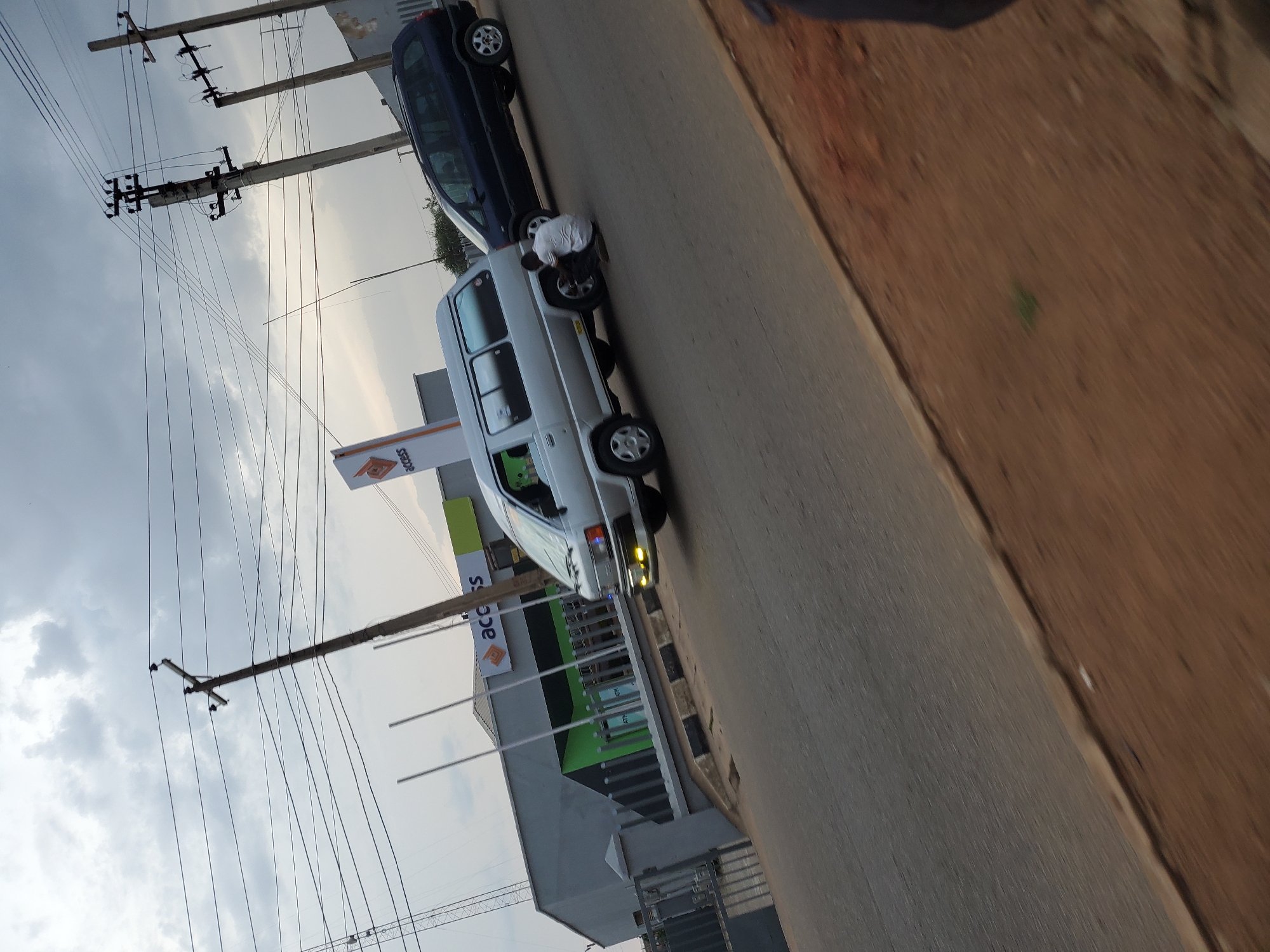 A medium shot captures a street scene in Bukuru, Nigeria, under a cloudy sky. In the foreground, a paved road stretches diagonally, with a dirt shoulder on the right. A silver van, possibly a Toyota Hiace, is parked on the left side of the road, facing away from the camera. A person in a white t-shirt and dark pants is crouched beside the rear passenger side tire of the van, appearing to be working on it. To the right of the silver van, a dark blue SUV is also parked, facing the same direction.
In the background, a low-rise building with a grey facade and green accents is visible, identified by an "Access" logo, suggesting it is a bank. Several utility poles with a network of electrical wires crisscross the sky above. The overall atmosphere suggests an ordinary day in a Nigerian city, with vehicles and infrastructure characteristic of the region.
