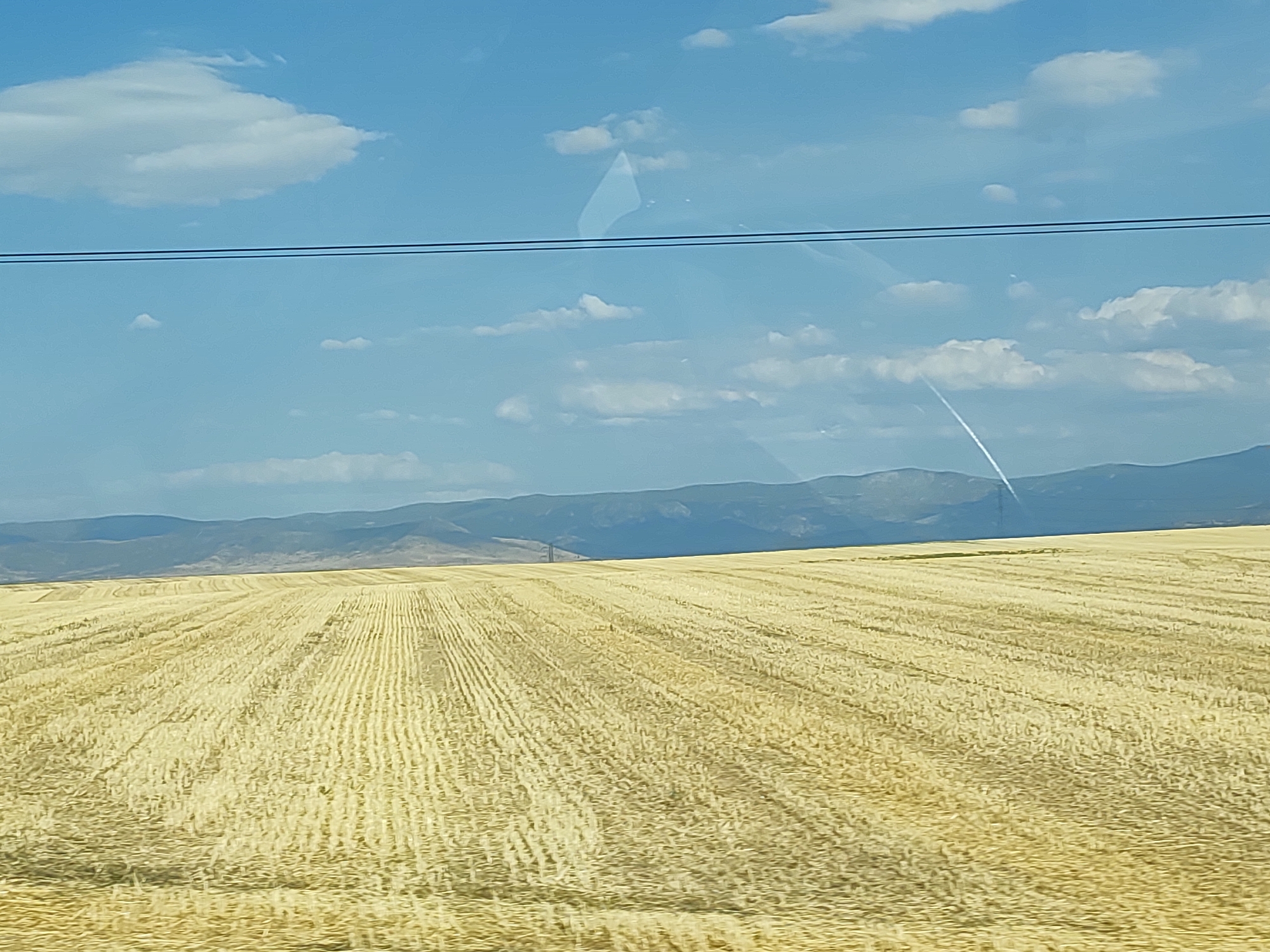 The image depicts a vast, harvested wheat field in Valverde del Majano, Spain, under a clear blue sky with scattered white clouds. The field displays distinct rows of stubble, indicating recent agricultural activity. In the background, rolling hills and mountains form a distant, muted silhouette against the horizon. Two black power lines traverse the upper portion of the frame, casting faint reflections on what appears to be a window. A thin, white contrail is visible diagonally across the sky. No people or specific structures are discernable within the field itself. The overall scene is one of rural tranquility and open, expansive agricultural land.
