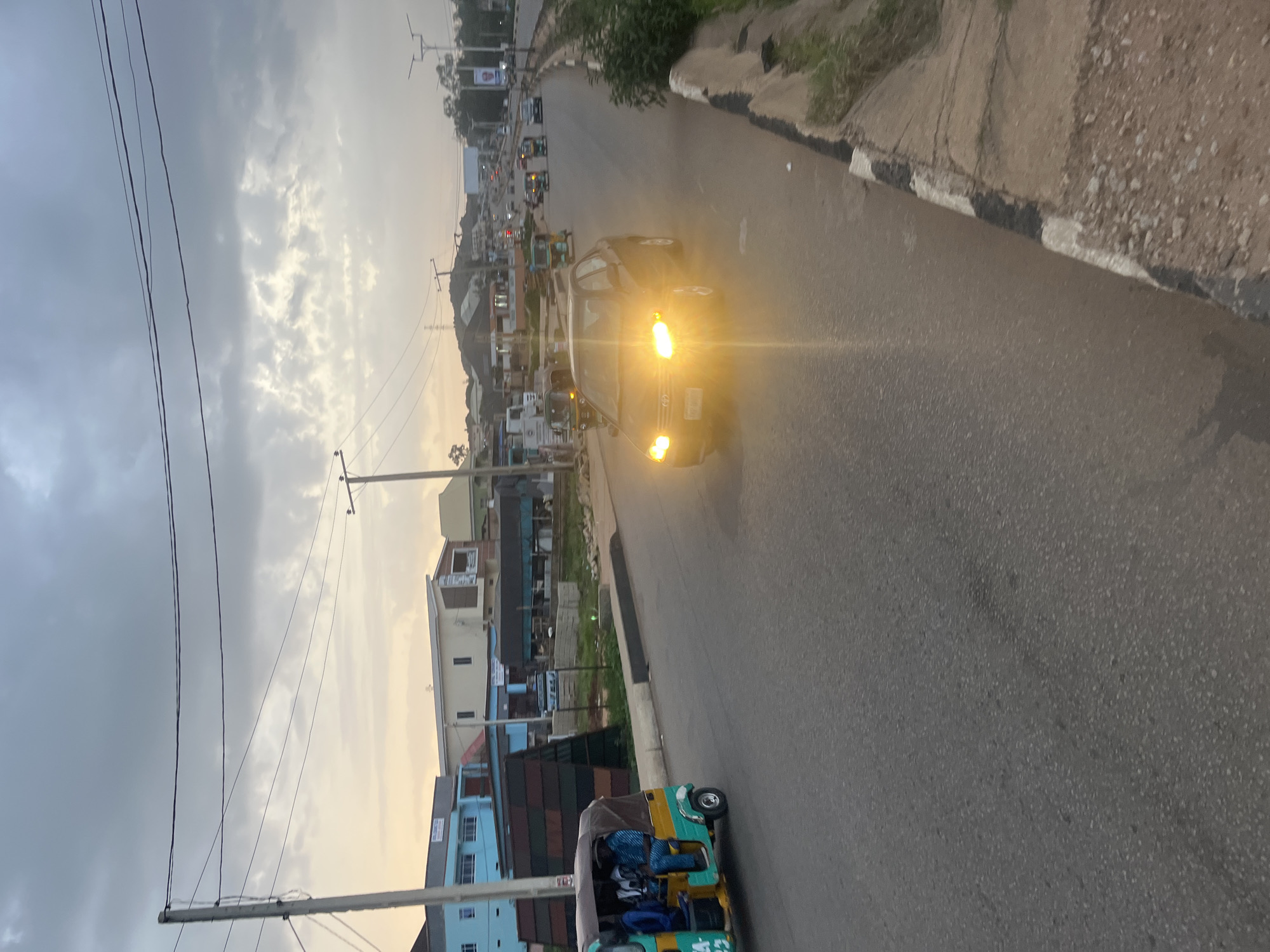 The image depicts a street scene in Gura Top, Nigeria, likely during dusk or overcast conditions. A dark-colored sedan with its headlights on is driving on the right side of the road, its bright beams creating a glare that obscures details of the vehicle and the road ahead. To the left, a yellow and green auto-rickshaw, also known as a Keke Napep, is moving in the same direction. This auto-rickshaw carries at least three passengers, including a driver. The surrounding environment features a paved road with a concrete curb on the right side. Beyond the curb, there is a patch of greenery and rough terrain. In the background, various buildings, including multi-story structures with blue and beige facades, are visible, suggesting an urban or semi-urban setting. Power lines and utility poles are prominent features of the overhead landscape. The sky is cloudy, with patches of lighter sky visible, indicating diffused sunlight. The overall impression is one of a typical day's traffic on a Nigerian road.