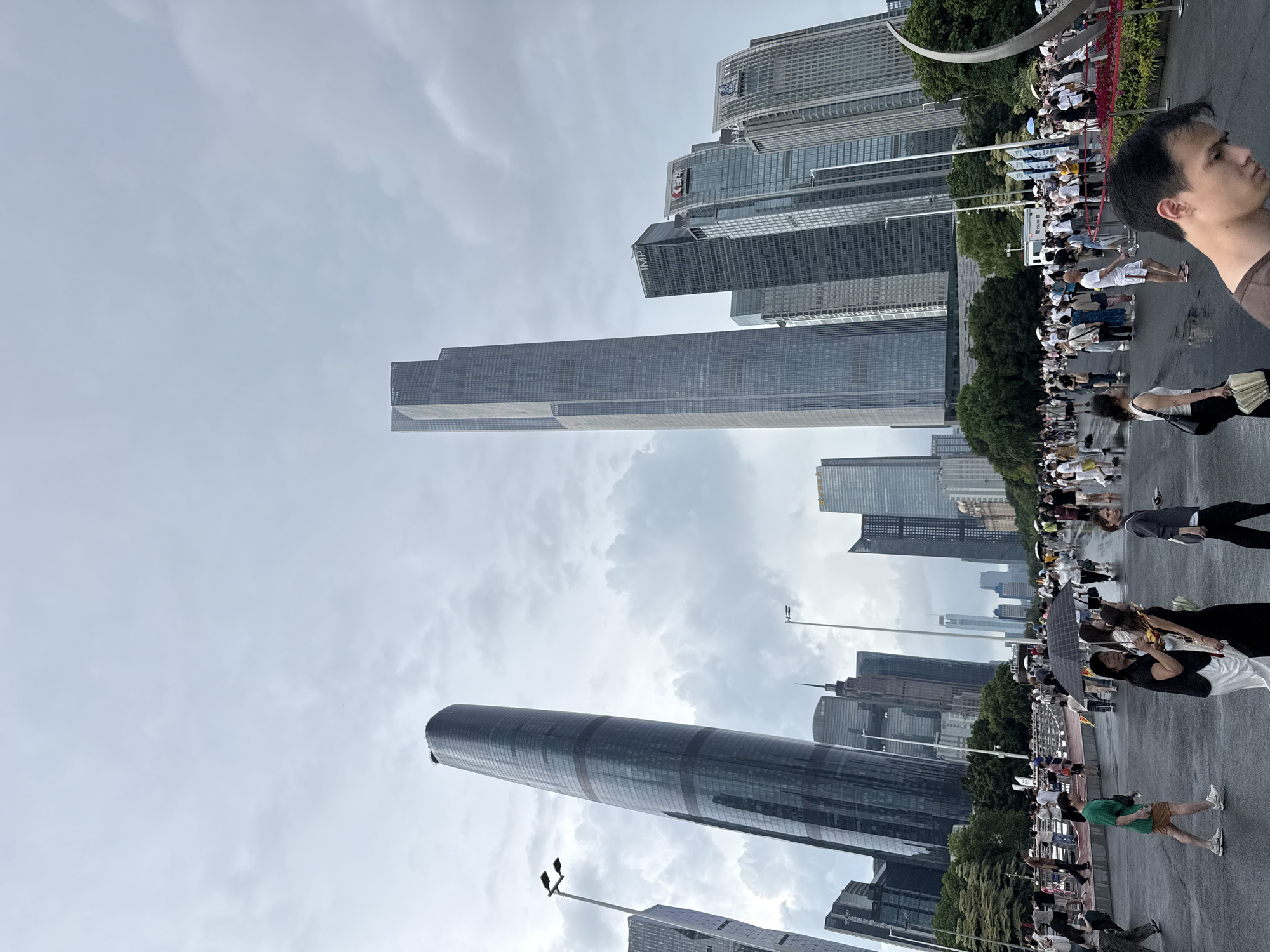 The image depicts a wide, wet public plaza in Tianhe District, China, with numerous people gathered. Dominating the background are several modern skyscrapers, including a distinctively curved, dark glass building on the left and a very tall, gray, rectangular skyscraper to its right. Other tall buildings with glass facades and some featuring corporate logos are also visible. The sky is overcast with gray clouds. In the foreground, the plaza surface is wet, reflecting the surrounding environment. A diverse crowd of people are walking, standing, and interacting. Some individuals are holding umbrellas, suggesting recent or ongoing rain. A few are carrying bags, and one person is holding a fan. The scene appears to be an urban public gathering space, possibly a promenade or a square, with a notable presence of contemporary architecture.