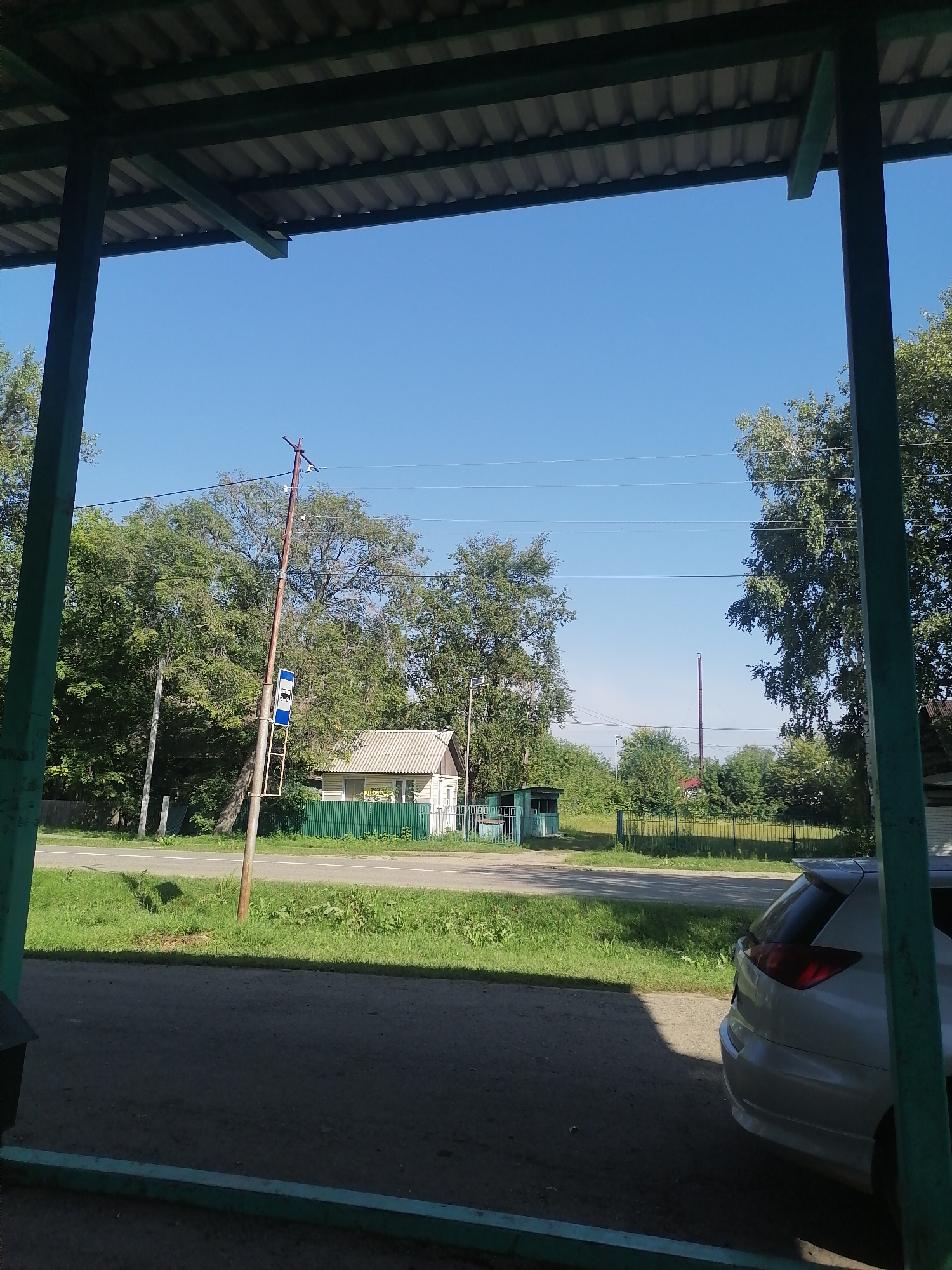 This image depicts a scene in Novopokrovka, Russia, viewed from under a covered structure. In the foreground, the ground is paved with asphalt, transitioning to a grassy area with a road beyond. A silver hatchback car is parked on the right side of the asphalt. The middle ground features a road with a grassy median, a bus stop sign on a pole, and a small building with a green fence in front. The background is dominated by lush green trees under a clear blue sky. Electrical wires are visible strung between poles. The overall impression is a rural or suburban setting on a sunny day.