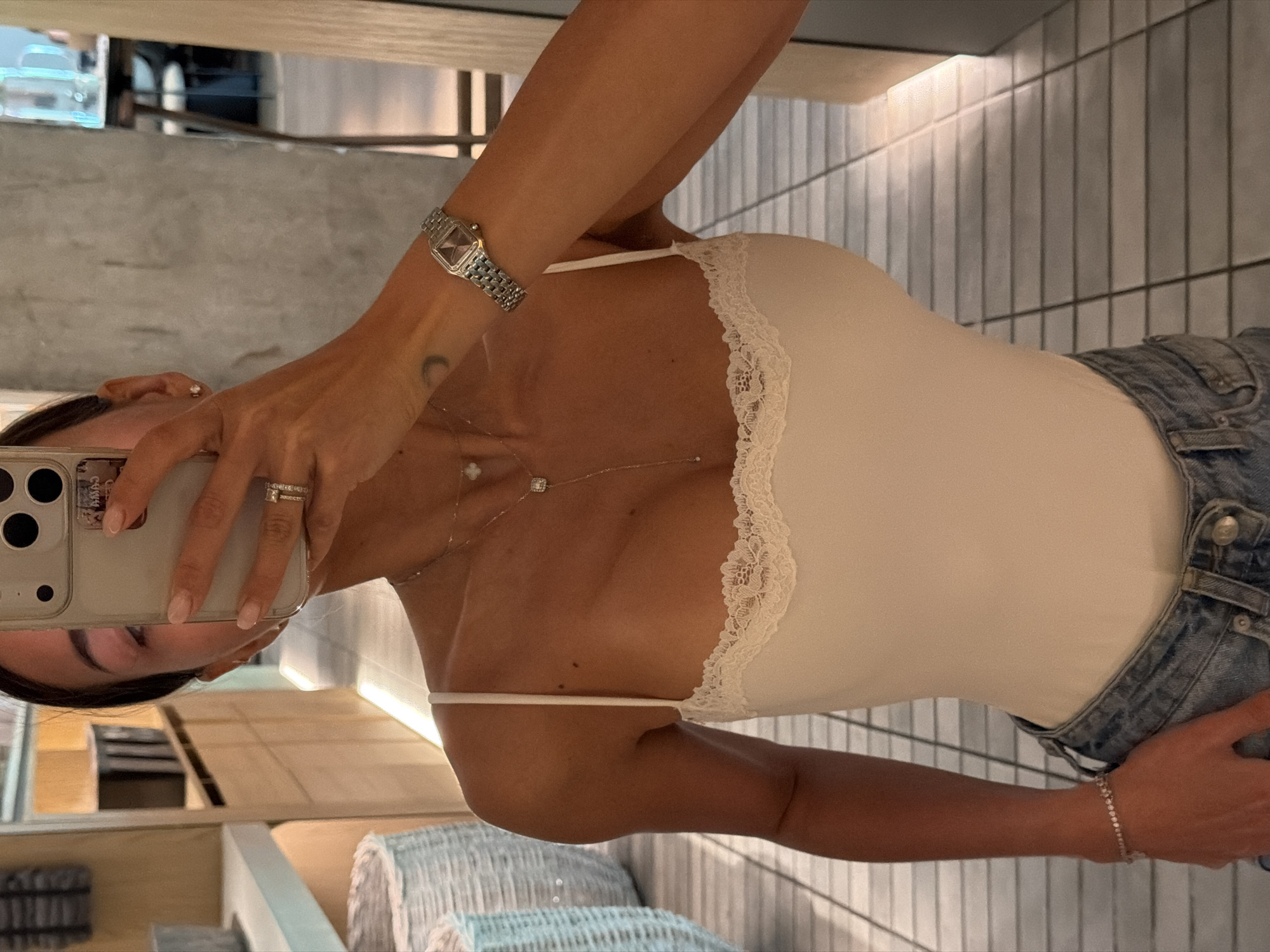 A woman is taking a selfie in what appears to be a bathroom or dressing room. She is wearing a white camisole with lace trim and denim jeans. She has on a silver wristwatch and rings on her fingers, and she is holding a smartphone to take the photo. There's a necklace visible around her neck. In the background, there are light-colored tiled walls and what appears to be a reflection of a room with furniture. There is no indication that the photo was taken in Krasnoyarsk, Russia.