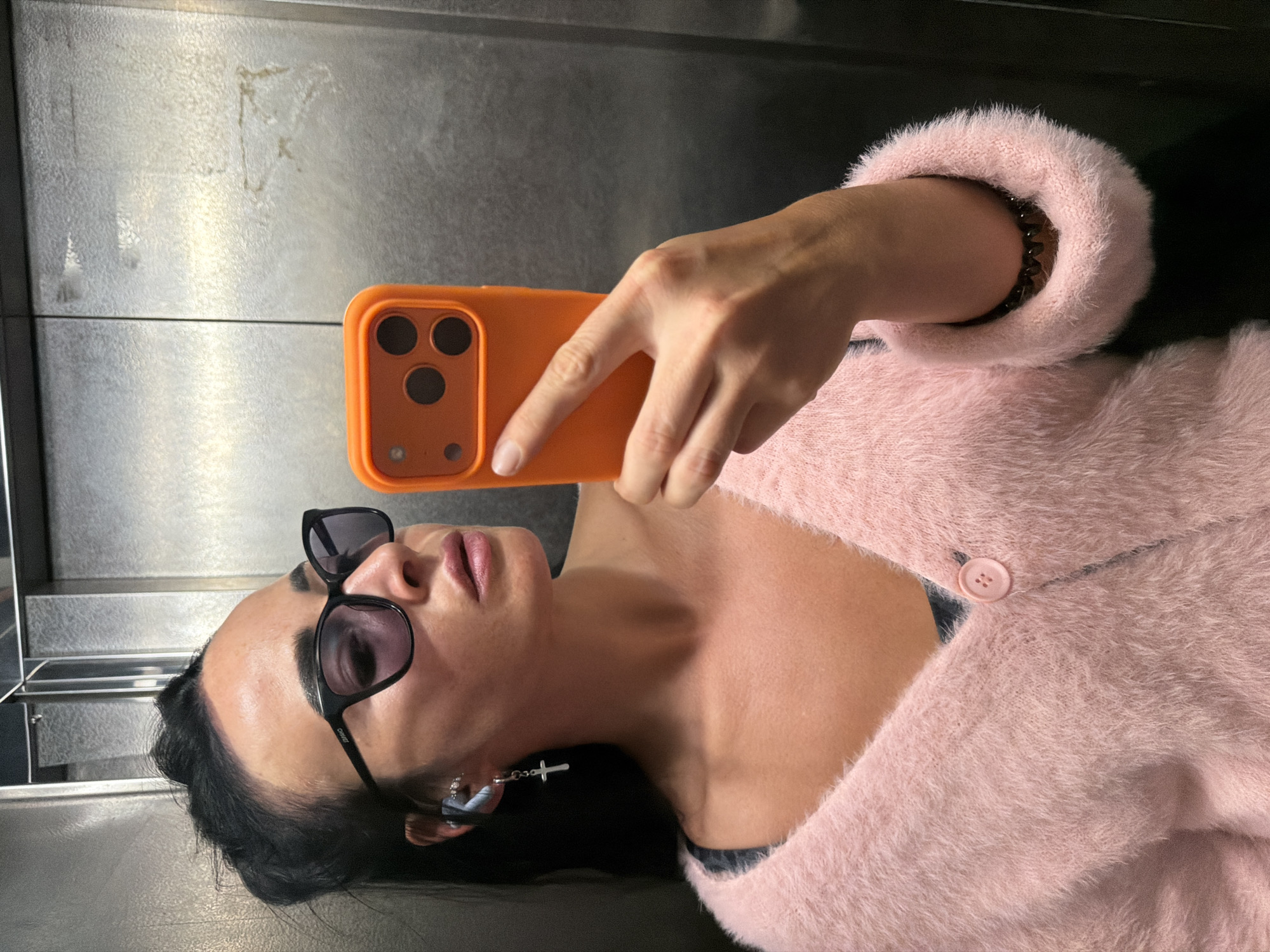 A woman is taking a selfie in an elevator. She is wearing a pink fluffy cardigan over a dark-colored top, black sunglasses, and a silver cross earring. She is also wearing an earpiece in her left ear and a bracelet on her right wrist. She is holding an orange phone with three camera lenses. The elevator appears to be made of brushed metal. The image does not contain enough information to determine the specific location within Saint Petersburg, Russia.