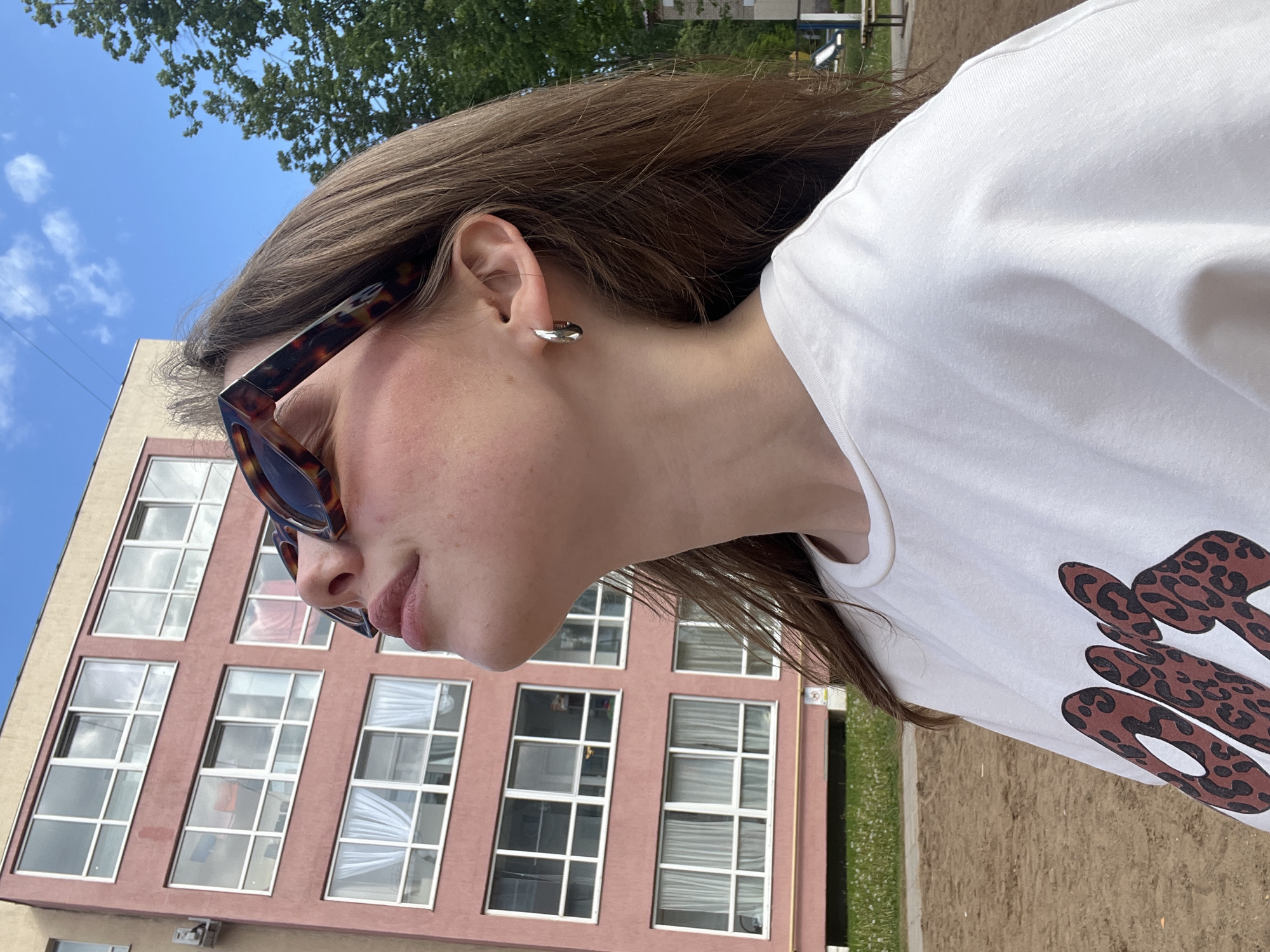 A side profile portrait captures a young woman outdoors in bright daylight. She is positioned facing left, with her head slightly tilted. Her long brown hair flows around her, and she wears stylish tortoiseshell-patterned sunglasses on her face. A small, simple silver hoop earring adorns her right earlobe. She is dressed in a white t-shirt that features a distinctive graphic: a partial word in a chunky, rounded, brown leopard-print font, possibly part of "GOOD" or a similar word.
The background prominently features a multi-story building painted in a faded reddish-pink or salmon hue, characterized by numerous large rectangular windows with white frames. Above the building, a clear blue sky with scattered white clouds is visible, and the tops of green trees can be seen in the distance. The ground in the lower right consists of light brown earth or sand mixed with patches of green grass. The bright and clear lighting indicates a sunny day, likely midday or afternoon. A slight natural flush or redness is noticeable on her cheek. The setting appears to be an urban or suburban environment, consistent with Chaikovsky, Russia.