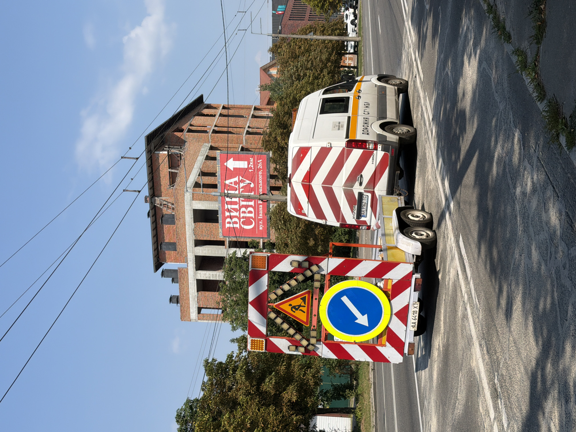 This is an eye-level, medium shot of a street scene in Kyiv, Ukraine, under a clear blue sky with scattered clouds. In the foreground, a construction trailer with a large red and white striped sign is positioned in the road. The sign features warning lights, a triangular road sign depicting a worker with a shovel, and a circular blue sign with a white arrow pointing left. The trailer's license plate reads "AA 6418 XT".
A white van with red and white chevron markings and the words "ДОРОЖНЯ СЛУЖБА" (Road Service) in orange lettering is parked to the right of the trailer. This van's license plate is "AA 9162 TC". In the background, a multi-story building under construction, made of red brick, dominates the skyline. Across the street, another building with a red roof and a modern facade is visible. Trees line the sides of the road. The road itself appears to have construction debris or dust scattered on it, particularly near the trailer. The image captures a moment in time related to road work or traffic management within the urban environment of Kyiv.