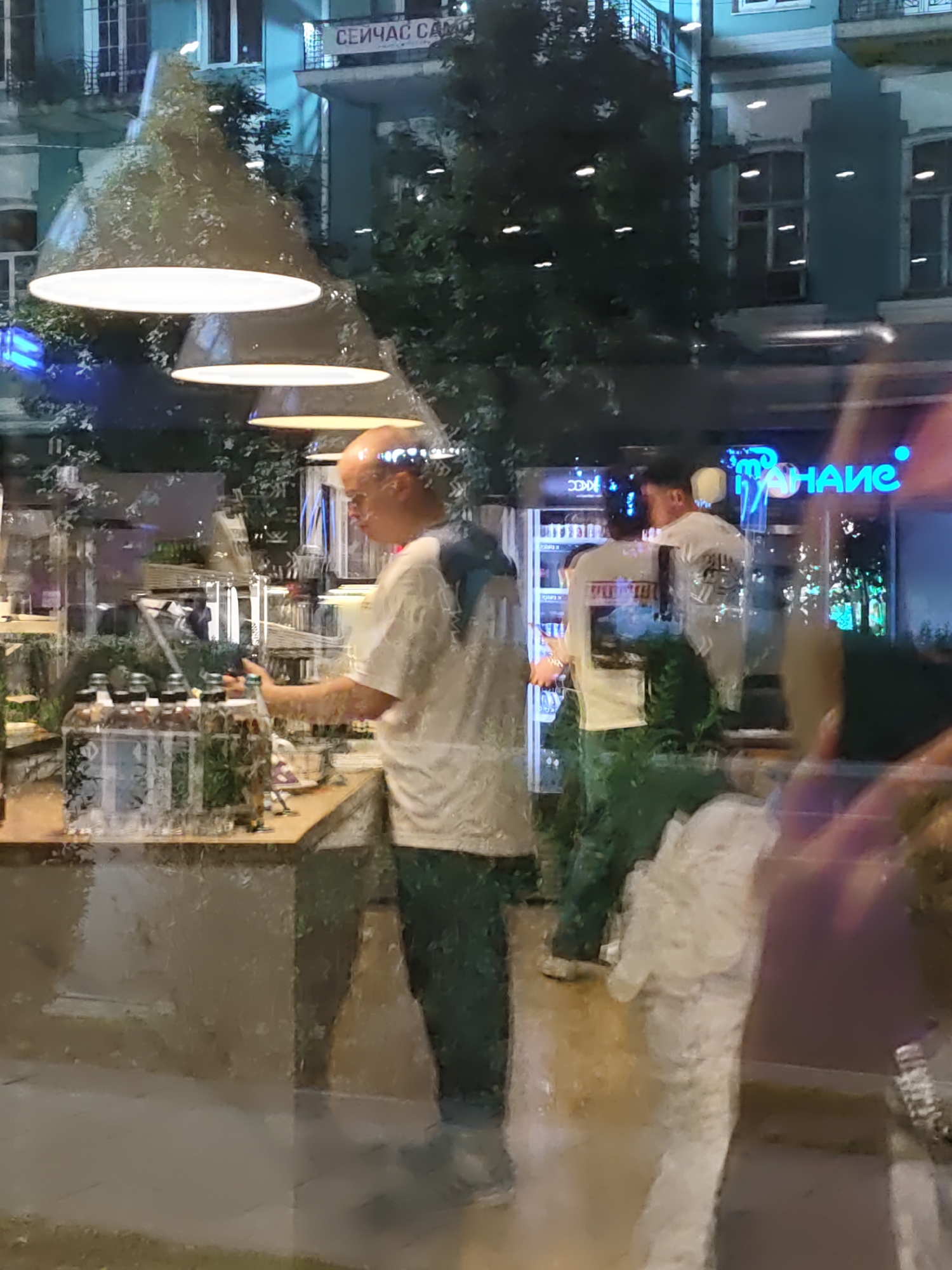 The image depicts a street-level view through a window, likely from a shop or cafe, into an interior space. In the foreground, a person, appearing to be a male with a shaved head, is standing behind a counter, looking down at items on the counter, possibly bottles. He is wearing a white t-shirt and dark green trousers. To the right, another person, also appearing to be male, wearing a white t-shirt with a graphic print and dark green trousers, is visible. He is standing near a refrigerated display unit with blue neon lighting. Behind the people, a bright blue neon sign with Cyrillic lettering is partially visible. The background reveals the exterior of a building with windows and balconies, and trees. The overall lighting is dim with interior lights and the neon sign providing illumination. The reflections on the window add a layer of visual complexity. This scene is located on Pushkinskaya Street in Rostov-on-Don, Russia.