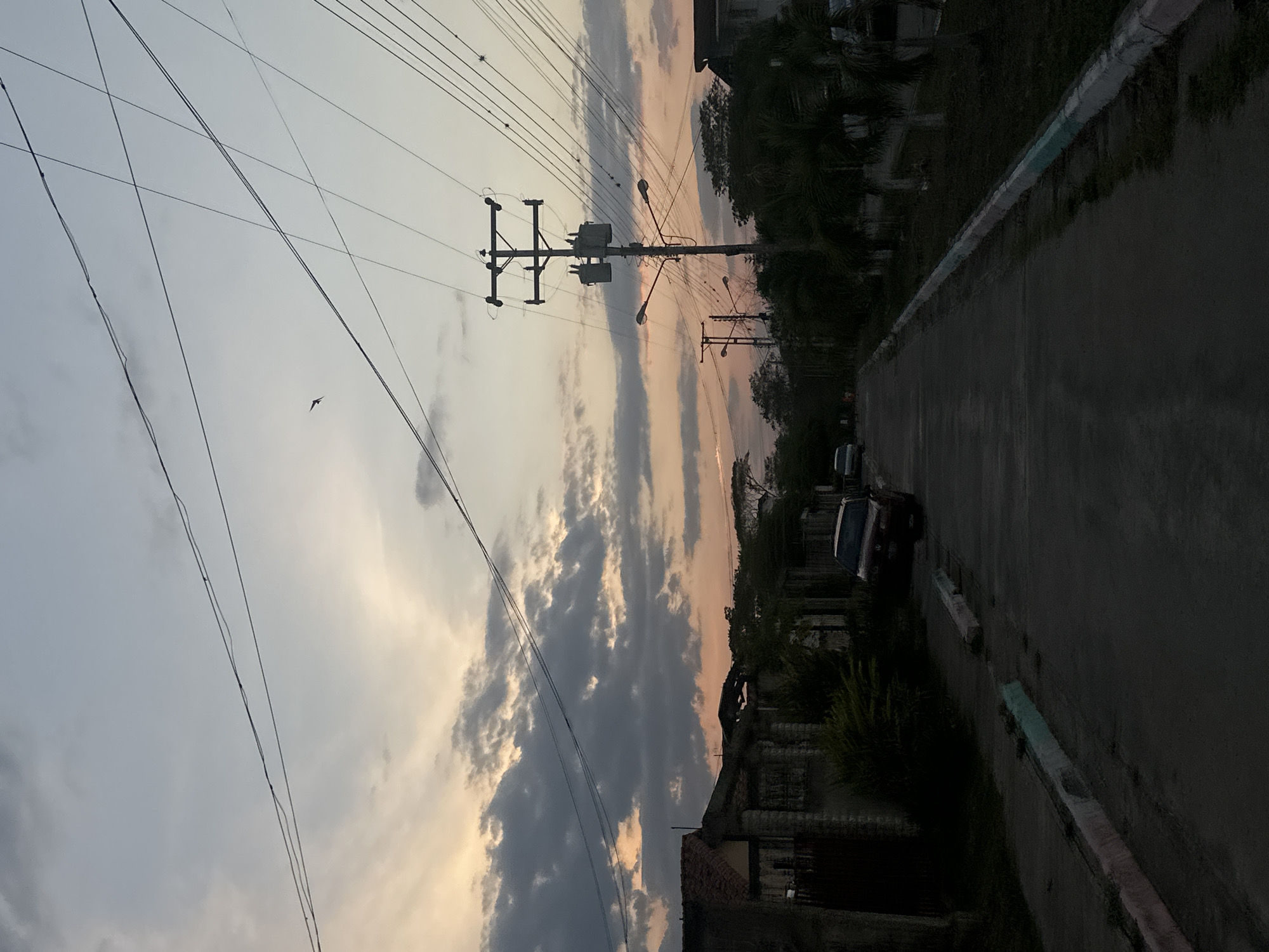 The image shows a street scene in Parroquia Ocumare, Venezuela, at dusk. A paved road runs through the scene, flanked by buildings and vegetation on the right. A dark-colored car is parked along the side of the road. Overhead, power lines crisscross the sky, connected to utility poles. The sky is filled with clouds illuminated by the setting sun, with colors ranging from gray to orange. A single bird is visible in the sky. The image appears to be taken at ground level, looking slightly upward.
