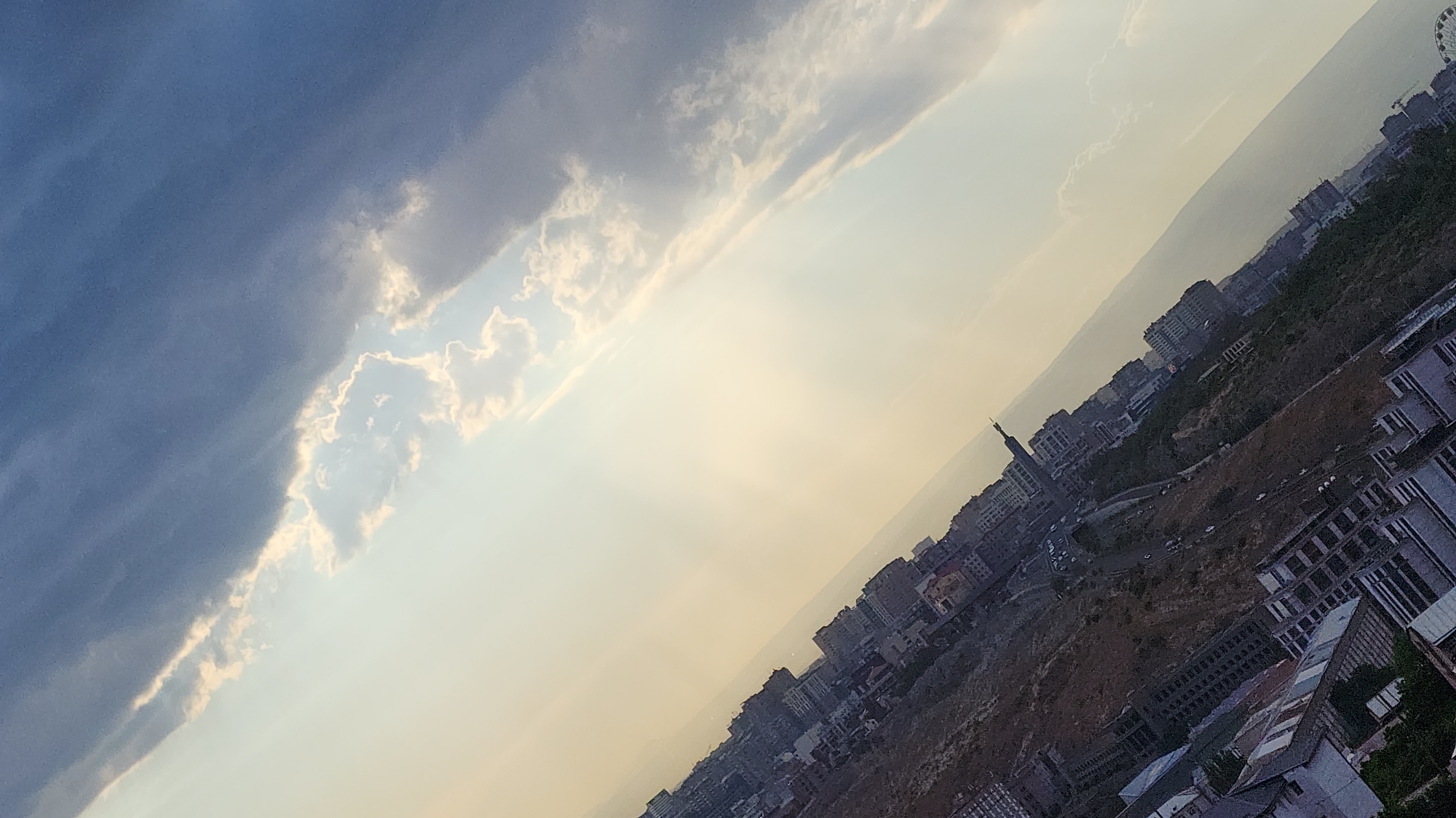 The image is an aerial shot of Yerevan, Armenia, taken during what appears to be late afternoon or early evening. The perspective is from a high vantage point, looking down and across a city landscape. The photo is tilted, with the sky occupying the upper left portion and the cityscape stretching diagonally across the lower right.
The sky is a dramatic mix of dark, moody clouds in the upper left, transitioning to a bright, hazy expanse of lighter clouds and clear sky as it moves towards the right. Rays of sunlight pierce through the clouds, creating a luminous, almost ethereal effect in the central part of the sky.
Below the sky, the city of Yerevan is visible, built on a hilly terrain. Numerous buildings of varying heights and architectural styles are scattered across the landscape, interspersed with patches of greenery and darker, uncultivated ground. In the middle ground, a prominent, slender tower with a spire stands out, hinting at a significant landmark. Roads with some vehicular traffic are discernible winding through the city. In the far right of the image, a large Ferris wheel is partially visible, suggesting an entertainment or recreational area within the city. The buildings in the foreground appear more densely packed and modern, with some showing unfinished construction.
The overall mood is serene, with the dramatic sky contrasting with the urban sprawl. The lighting suggests a transition period of the day, perhaps sunset, casting a warm glow over parts of the scene. No discernible text is visible in the image.