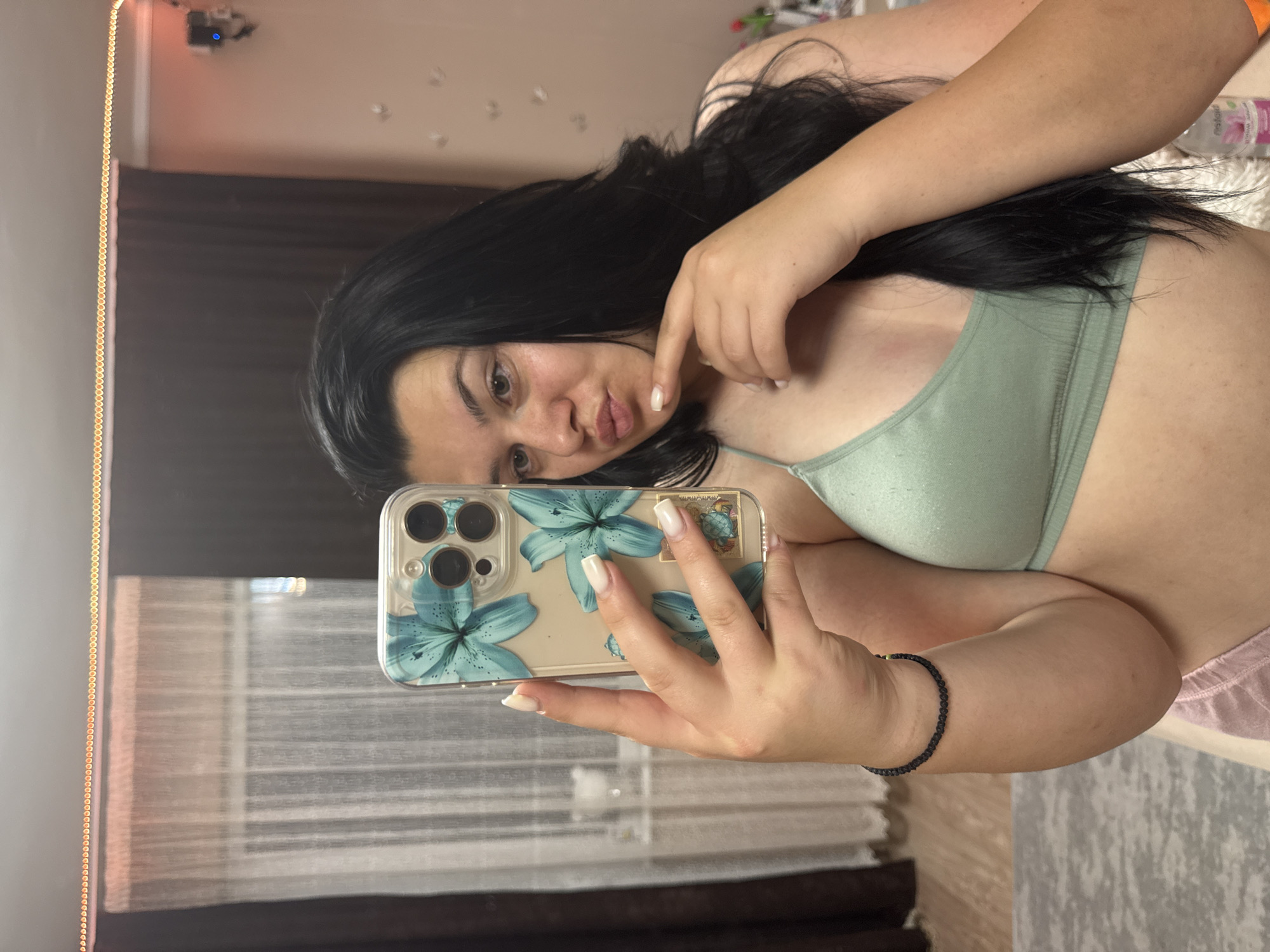 A young woman with long, dark hair and light brown eyes is taking a selfie in a room. She is wearing a light green bralette and light pink shorts. Her left hand is holding up a smartphone with a blue floral case. Her right index finger is touching her chin. The background of the image includes a sheer white curtain on the left, a dark brown curtain behind her, and a wall with decorative lights and a small speaker on the right. The flooring appears to be wood with a grey rug. The setting is likely a bedroom. There are no identifiable landmarks that place the image specifically in Rogosh, Bulgaria.