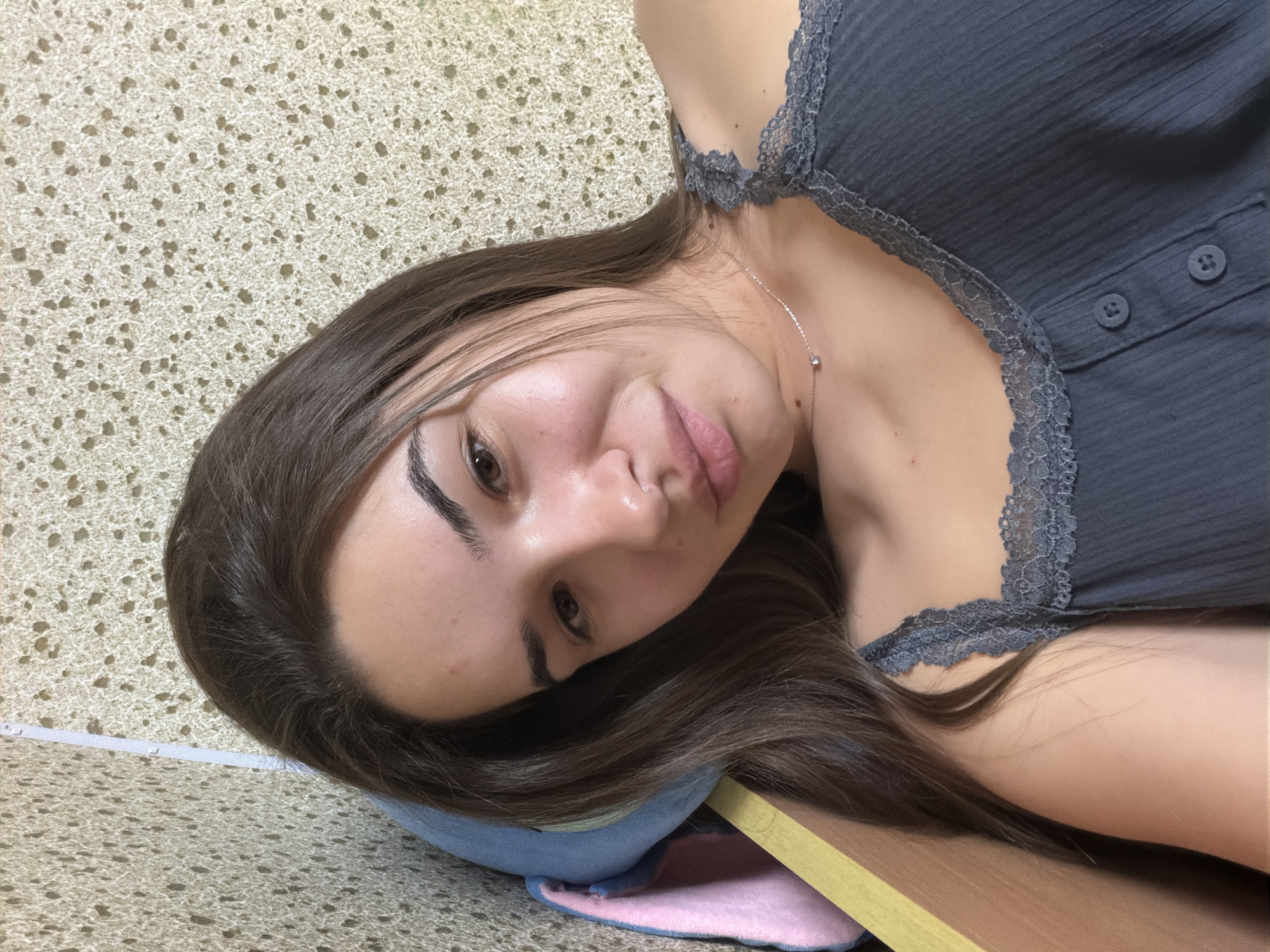 A young woman with long brown hair and light skin is lying down, her head resting on a blue pillow that is partially covering a pink pillow. She is wearing a gray tank top with lace trim. A silver necklace with a small pendant is visible on her neck. The surface she is lying on appears to be a desk or table with a light brown, speckled surface. A portion of what seems to be a measuring tape is visible near her head. The background is a light brown, speckled surface, possibly a wall or floor.
There is no location information present in the image to associate it with Sofia, Bulgaria.
