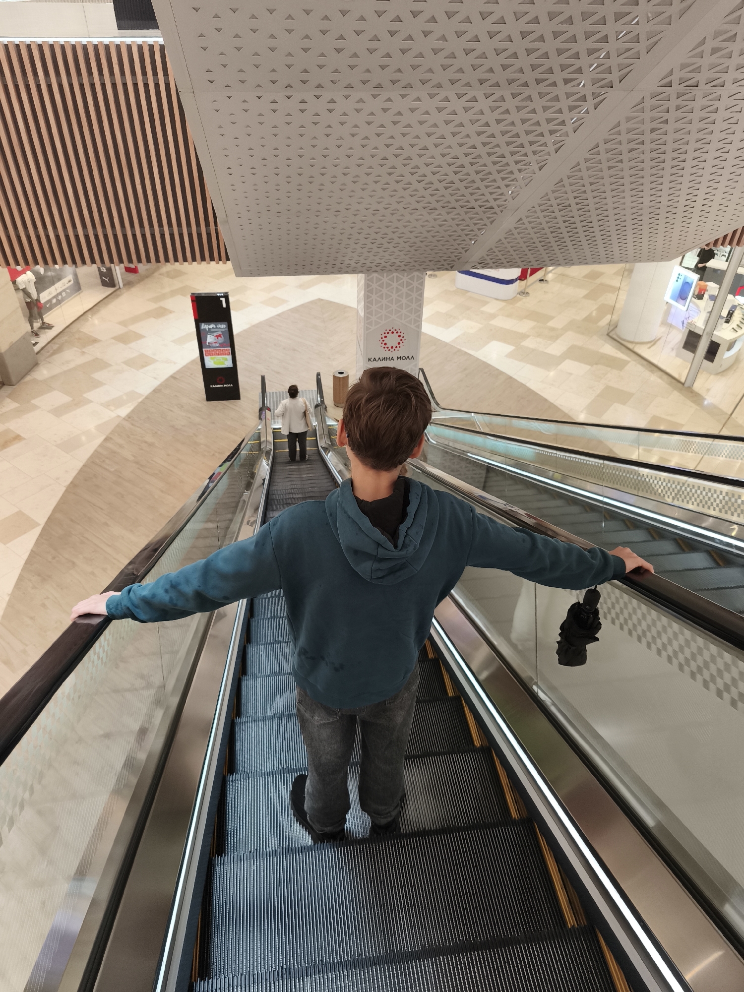 The image depicts a young person, seen from behind, riding an escalator downwards in a modern, multi-level shopping mall. The individual, wearing a teal hooded sweatshirt and dark jeans, has their arms outstretched, holding onto the escalator handrails. A black umbrella is hanging from their right hand. In the background, other shoppers are visible, as well as the architecture of the mall, including wood-paneled walls, a perforated ceiling, and illuminated displays. The floor is tiled in a light color with a patterned design. A sign with Cyrillic text, "КАЛИНА МОЛЛ," is visible, indicating the location as Kalina Mall in Vladivostok, Russia. The lighting is bright and even, typical of an indoor commercial space.