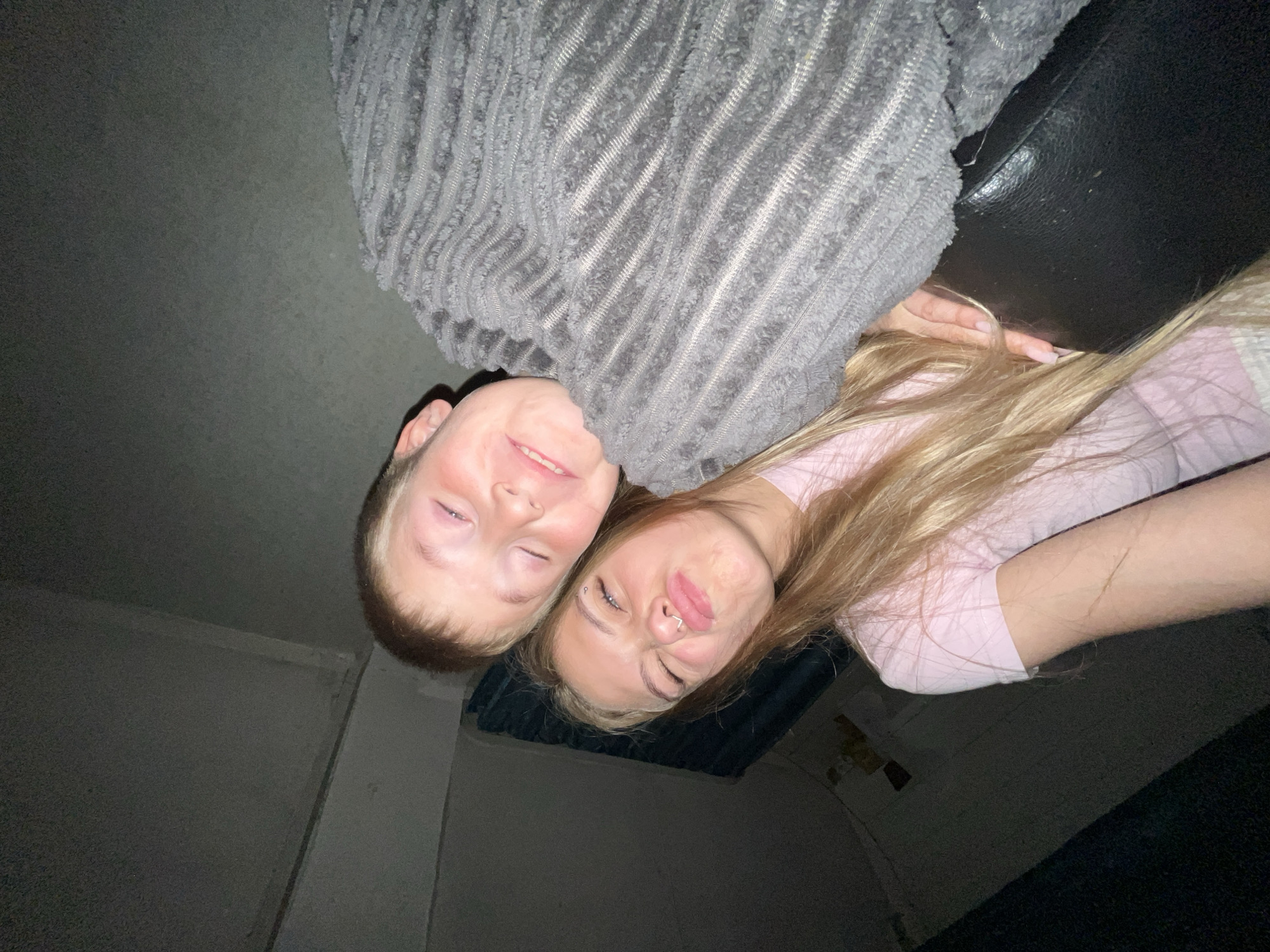 Two young people, a boy and a girl, are posing for a selfie in a dimly lit room. The boy, with short, light brown hair, is leaning his head against the girl's. He has his eyes closed and is smiling. The girl, with long, blonde hair, is looking at the camera with her eyes squinted and her lips pursed. She is wearing a pink t-shirt and has a nose ring. They are both covered by a textured grey blanket. The background is dark and indistinct, with some horizontal lines visible on the wall. The image appears to be taken in an indoor setting, possibly a bedroom or living room. The location in the city of Фоки, Russia is not discernible from the image.