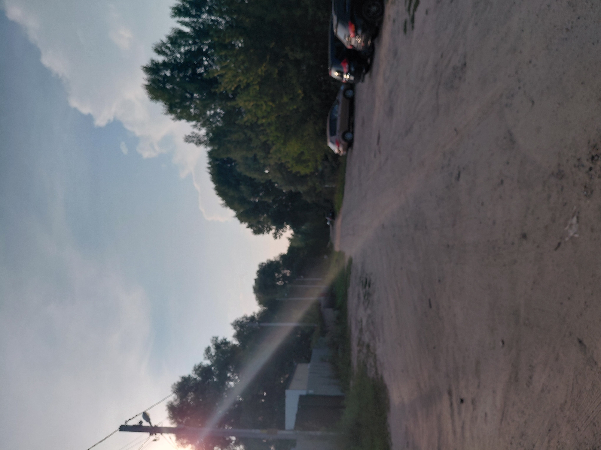 The image depicts a dusty, unpaved road in Novosibirsk, Russia, bordered by trees and a fence. The sun is low in the sky, casting long shadows and creating lens flares from a nearby utility pole. Several cars are parked along the side of the road, including a light-colored sedan and two dark SUVs. The scene appears to be a rural or suburban area, possibly on the outskirts of the city. The atmosphere is tranquil, with no people visible in the frame.