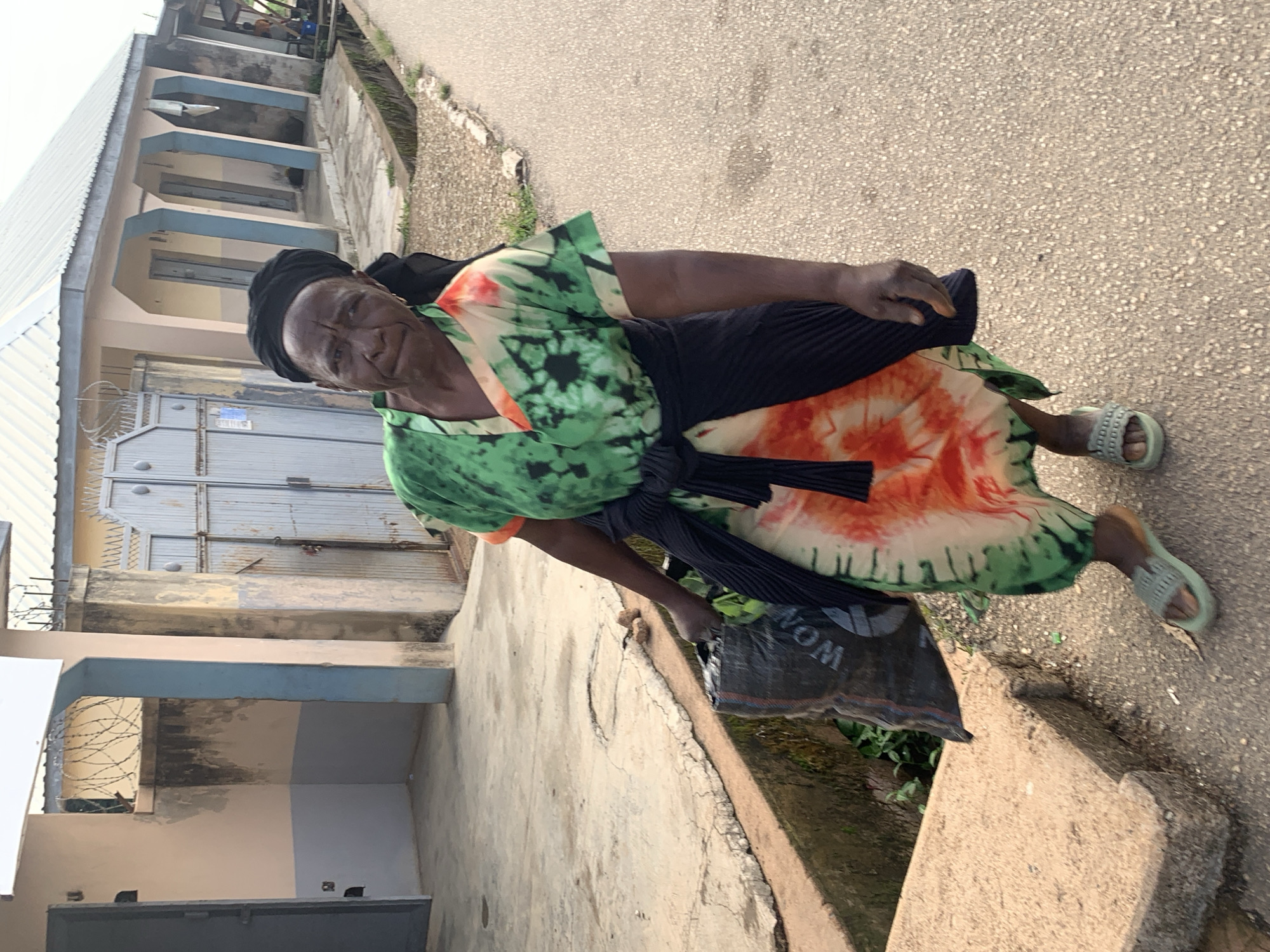 An elderly African woman walks along a paved road in Jos, Nigeria, carrying a black plastic bag filled with produce. She is wearing a green and orange tie-dye dress and a black headscarf. A black garment is tied around her waist. She wears light-colored sandals. The road is bordered by a concrete drainage channel and a sidewalk. Buildings with corrugated metal roofs are visible in the background. The scene is captured from a slightly low angle.