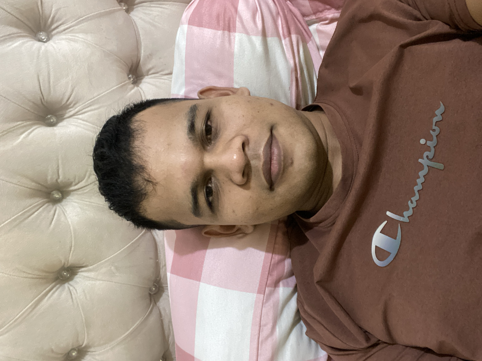 A man is lying on a bed, looking at the camera. He has dark hair and brown eyes, and is wearing a brown t-shirt with the Champion logo. The bed has a tufted headboard and a pink and white checkered pillow. The image appears to have been taken in Lambaro Kueh, Indonesia.