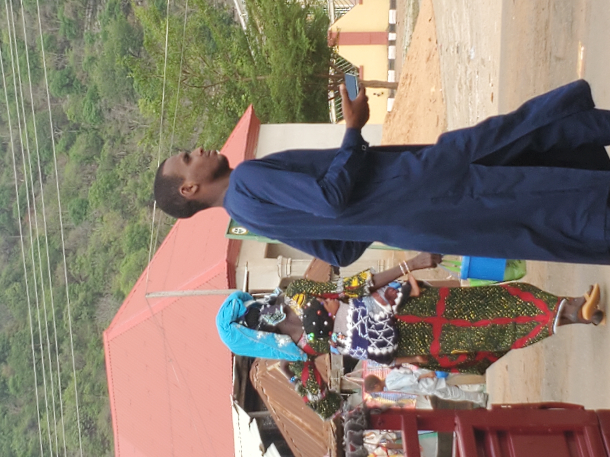 In Mararaba, Nigeria, an individual in a blue garment looks at a smartphone while standing on a street. Next to them is a woman in colorful traditional attire and blue headwrap. Nearby is a building with a red roof and another structure with a tan and reddish brown exterior. A forested hillside is visible in the background. A red metal structure is in the foreground.