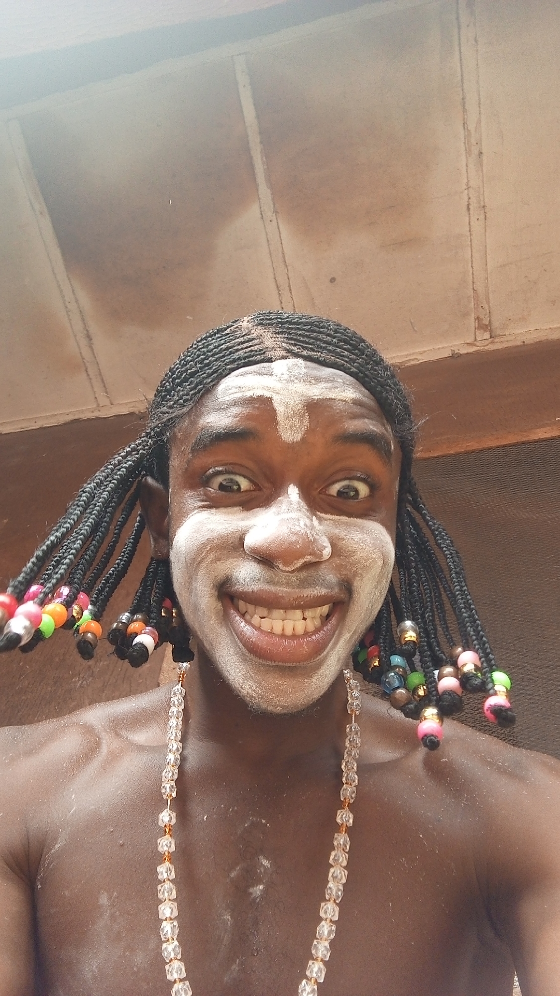 A close-up, wide-angle, low-angle shot shows a dark-skinned person with black braided hair decorated with colorful beads. The person has white powder or paint on their face, including a cross on their forehead. They are grinning widely, exposing their teeth. They wear a beaded necklace. The background shows a ceiling with visible seams, suggesting an indoor setting. The location is Onitsha, Nigeria. The person appears to be posing or performing.