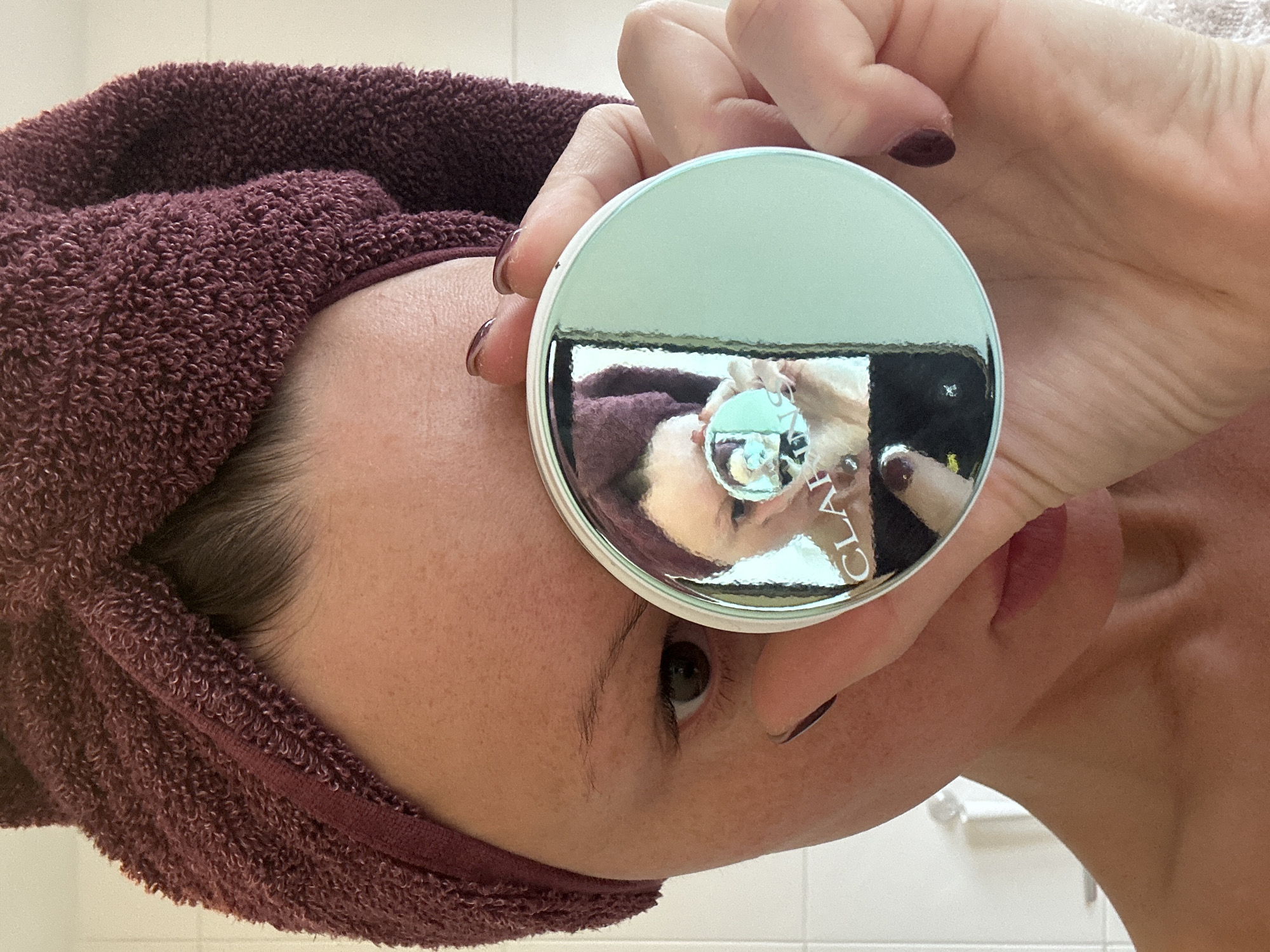 A person with a burgundy towel wrapped around their head is holding up a mirrored container in front of their face. The container is silver-green with a silver rim. The reflection in the container shows the person holding the container, the burgundy towel, and part of a white wall. The person has dark painted fingernails. The location cannot be determined from this image. Tunbridge Wells, United Kingdom is not identifiable.