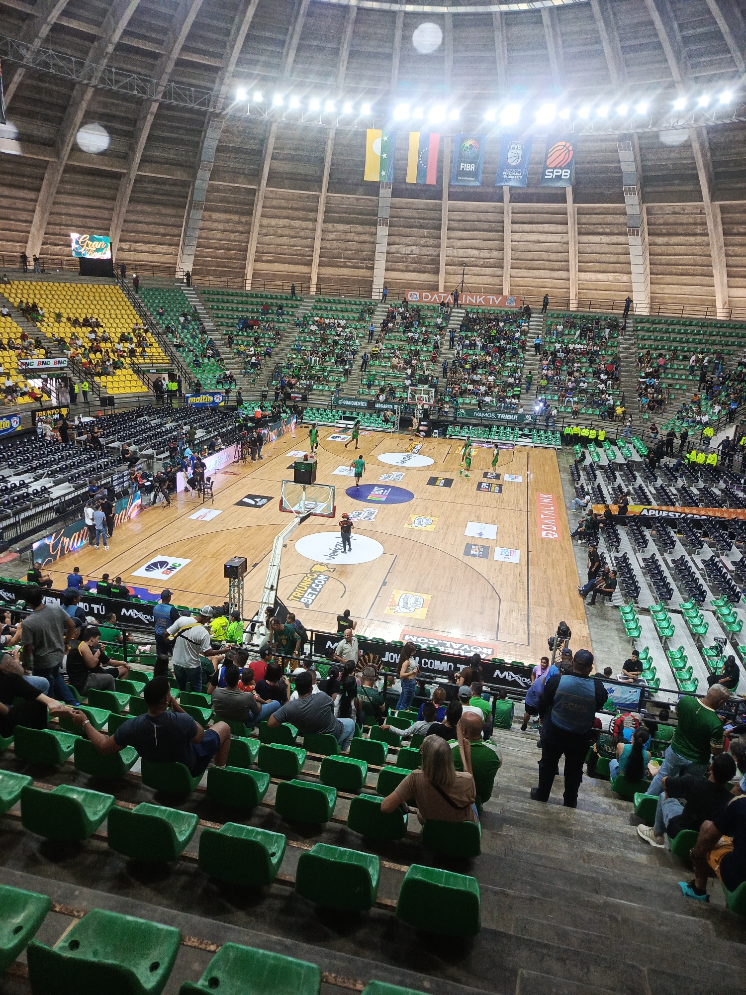 This is a high-angle, medium shot of a basketball game in progress within an indoor arena. The arena features tiered green and yellow seating, largely occupied by spectators. The court below is made of polished wood with various sponsor logos and markings. Several players in green uniforms are visible on the court, engaged in the game. Spectators are seated in the stands, some looking towards the court, others interacting amongst themselves. The arena's ceiling is a distinctive domed structure with exposed beams and industrial lighting. Banners hang from the upper structure, displaying flags and logos, including FIBA and SPB. In the foreground, spectators are seated in green chairs on concrete steps. The overall scene depicts a sporting event with a significant crowd in a large indoor venue. The specific location in the city of La Asuncion, Venezuela, is not explicitly identifiable from the image alone, although the presence of Venezuelan flags suggests a connection to the country.