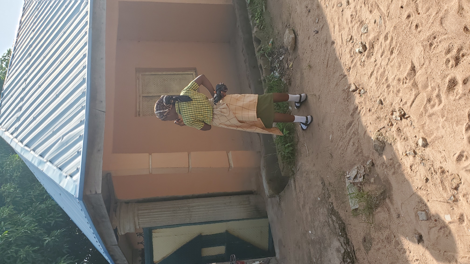 A young female stands on a dirt path outside a single-story, peach-colored building with a corrugated metal roof. She is facing away from the camera, wearing a yellow and black checkered short-sleeved shirt, a green skirt, a patterned headscarf, white socks, and sandals. A light-colored, checkered fabric is wrapped around her waist and draped over her skirt. She holds a dark object in her right hand. The building has a doorway on the left with a dark metal gate and a large, fluted column beside it. A window is visible on the right side of the building. The ground is covered in sand and scattered debris, with some sparse green vegetation growing in patches. The scene appears to be in an outdoor, possibly rural, environment on a sunny day. The location is identified as Jalingo, Nigeria.