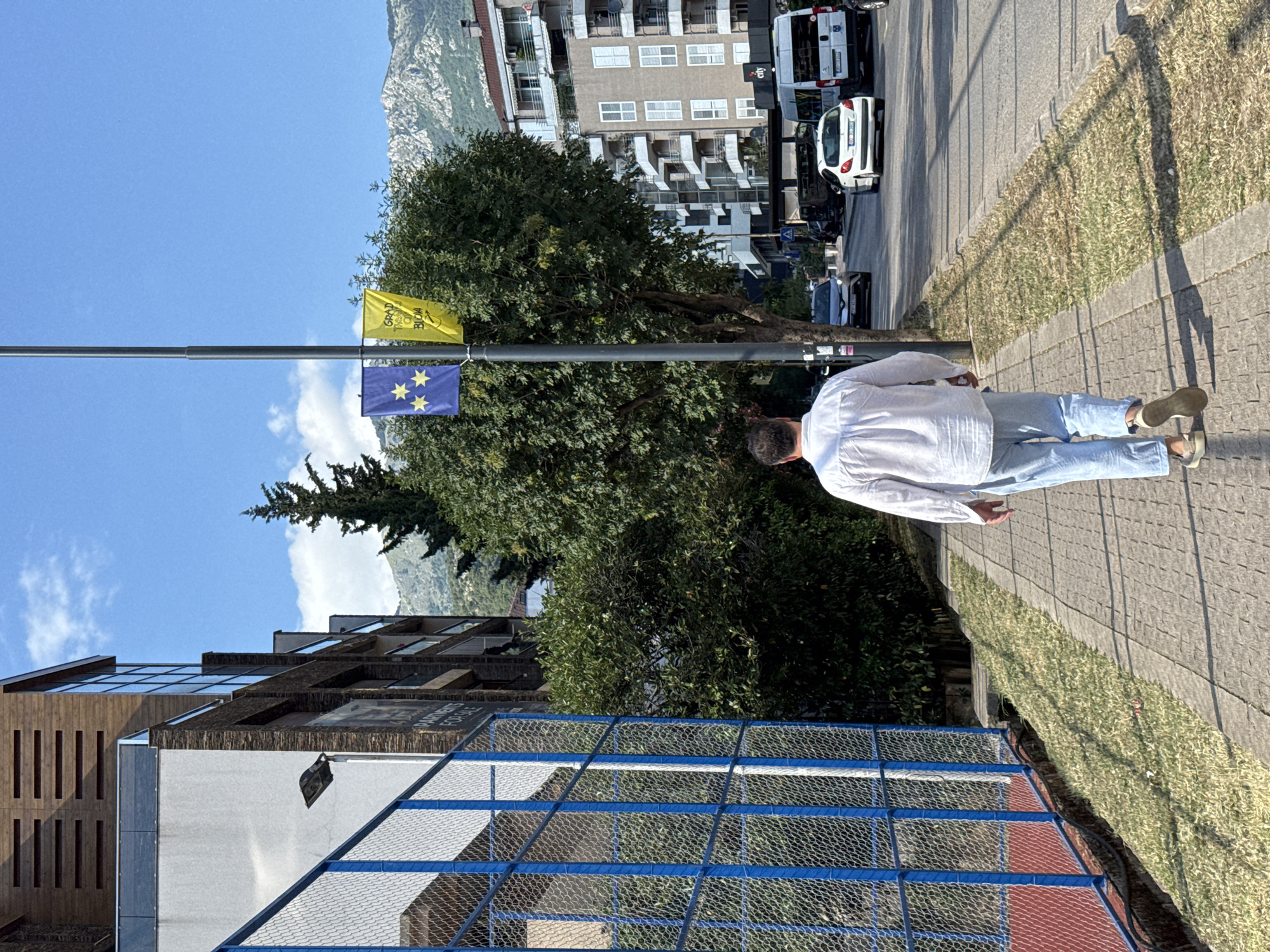 A person in a white shirt and light blue jeans walks along a sidewalk lined with grass. The person is facing away from the camera, and their right arm is extended downwards, with their hand slightly open. The scene appears to be a bright, sunny day, indicated by the clear blue sky and strong shadows.
In the background, there are several buildings, including a modern-looking structure with wooden accents and glass windows, and a taller building with multiple balconies. Across the street, there are more buildings and parked vehicles, including a white van and a white car. A street lamp with two flags is visible. One flag is yellow with dark text, and the other is blue with stars, resembling the European Union flag. Trees and vegetation are interspersed throughout the scene.
The image is oriented vertically, with the ground and sidewalk running from the bottom left to the top right of the frame. The overall setting suggests an urban or semi-urban environment, possibly a promenade or street in a town or city. The person's posture and relaxed clothing imply a casual stroll. The presence of the European Union flag could indicate a location in a country with ties to the EU or a pro-EU sentiment. There is a fence with blue metal framing and wire mesh in the foreground, suggesting a sports court or similar facility. The text on the yellow flag is partially visible and appears to be in a foreign language.