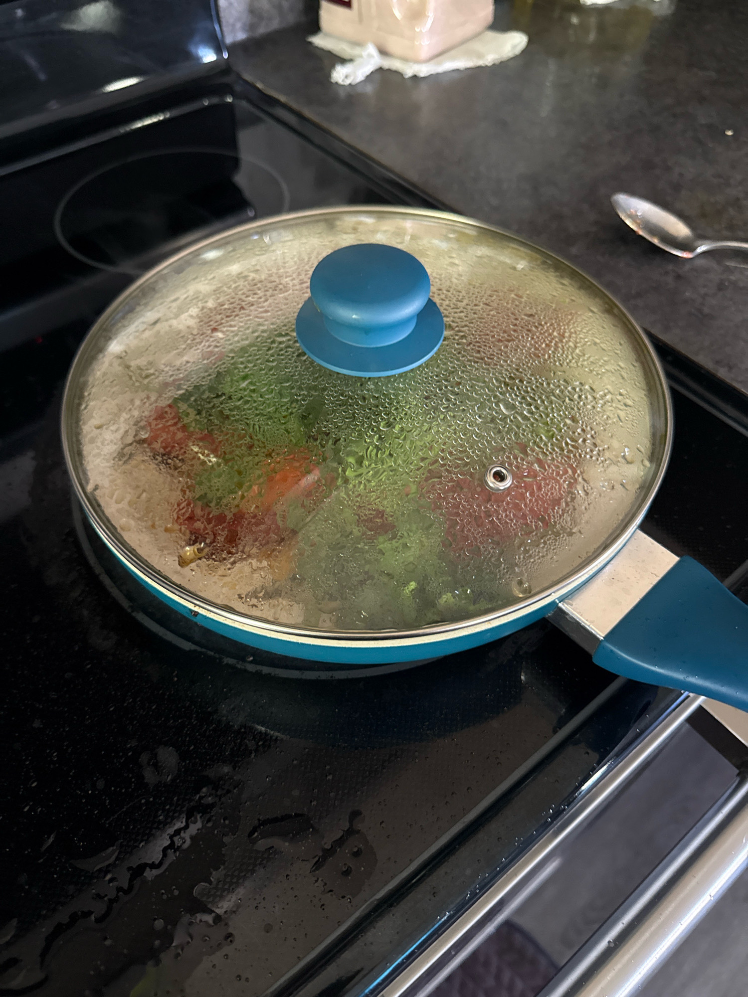 An overhead shot shows a teal frying pan with a glass lid on a black stovetop. Condensation is visible on the inside of the lid, obscuring the contents of the pan, which appear to be vegetables, possibly tomatoes and greens. The handle of the pan is also teal. A silver spoon rests on the stovetop to the right of the pan. The stovetop has a glossy black surface with circular heating elements. Water droplets are scattered across the stovetop. The background shows a dark countertop and a white cloth. This image was taken in Mora, United States of America.
