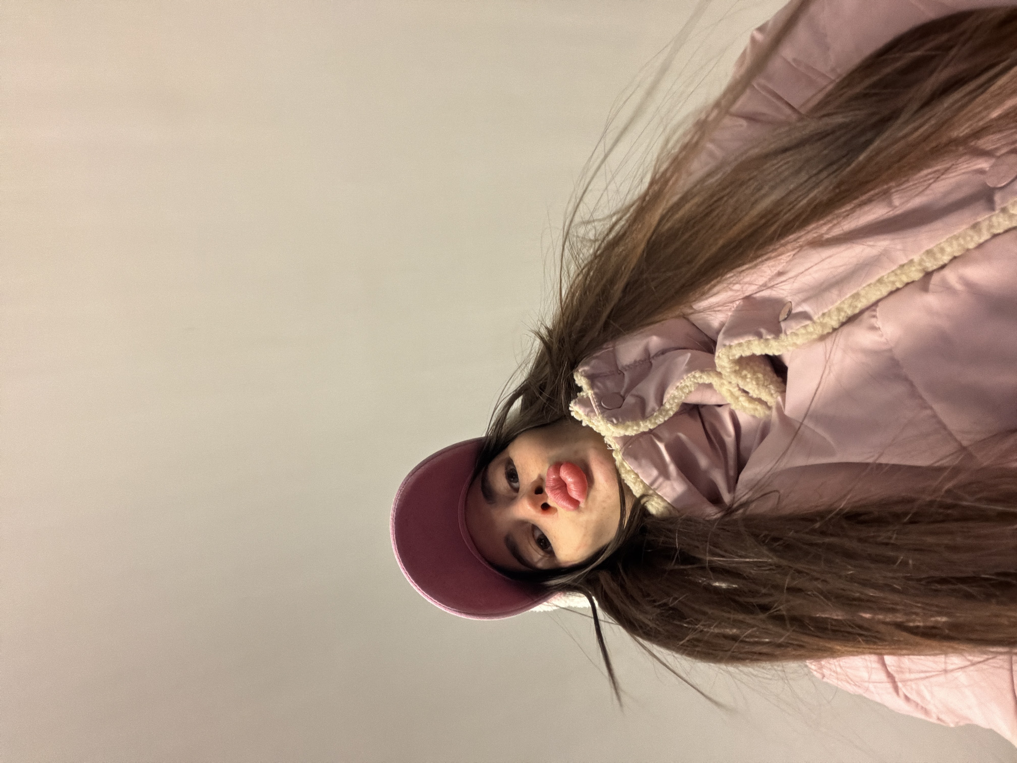A young woman wearing a pink baseball cap and a pink jacket with a cream-colored trim is seen from a low angle, looking directly at the camera with her lips puckered. The background is a neutral gray or white color. Her long, dark hair is spread around her head and shoulders. There are no visual indicators to definitively place this photo in Saint Petersburg, Russia.
