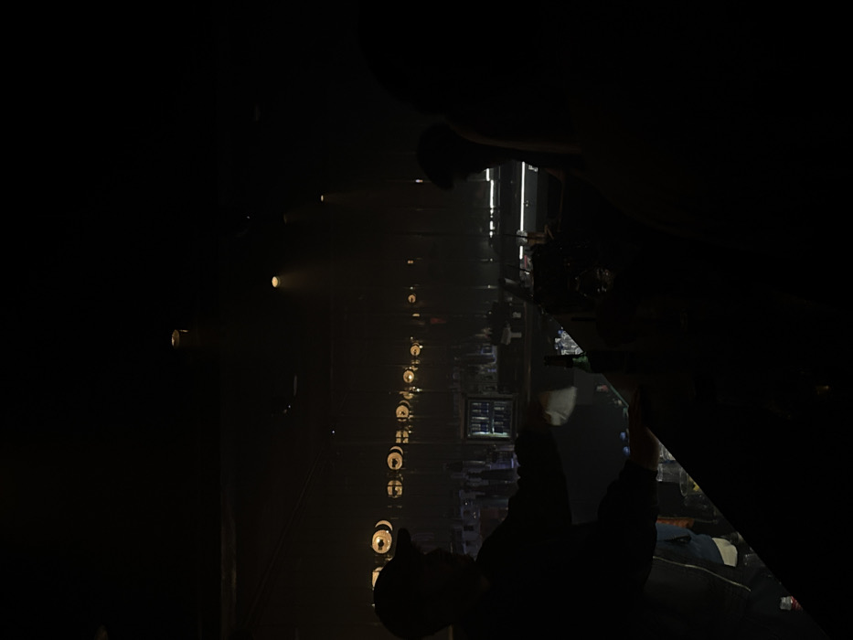 This image depicts the dimly lit interior of a nightlife establishment, likely a bar or club, located in Tel-Aviv, Israel. The specific venue name is not discernible from visual evidence. The scene is characterized by extremely low ambient light, with targeted illumination from overhead spotlights and a horizontal array of approximately 10-12 circular fixtures across the back wall. Visible haze or smoke interacts with the light beams. In the left foreground, a silhouetted male figure, wearing a dark cap and jacket, stands behind a bar counter. He is actively wiping the counter with a white cloth or napkin. On the right, a darker silhouette of a patron's head and shoulders is visible, facing the bar. The bar counter contains various bottles, including a prominent green bottle, and glassware. Behind the counter, a back bar displays shelves of bottles and an illuminated beverage cooler. Several indistinct figures are faintly visible in the deeper background, indicating other individuals present in the establishment.