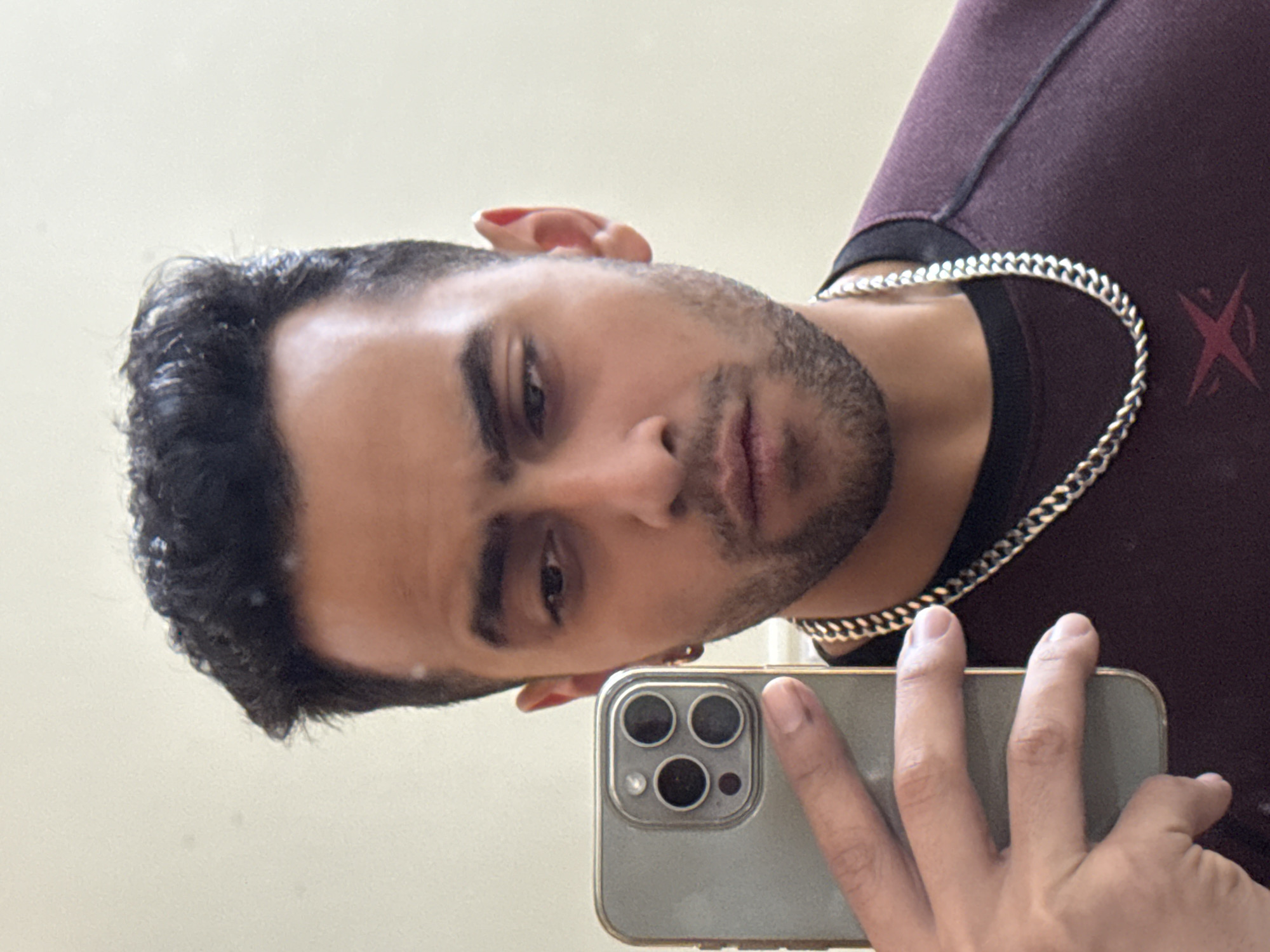 A man is shown in a selfie. He is holding a smartphone with a triple-lens camera. He has short, dark hair and a short beard. He is wearing a dark maroon shirt with a black collar, and a silver chain. He has a small earring in his left ear. The background is a plain, off-white wall. The location is not discernible from the image.