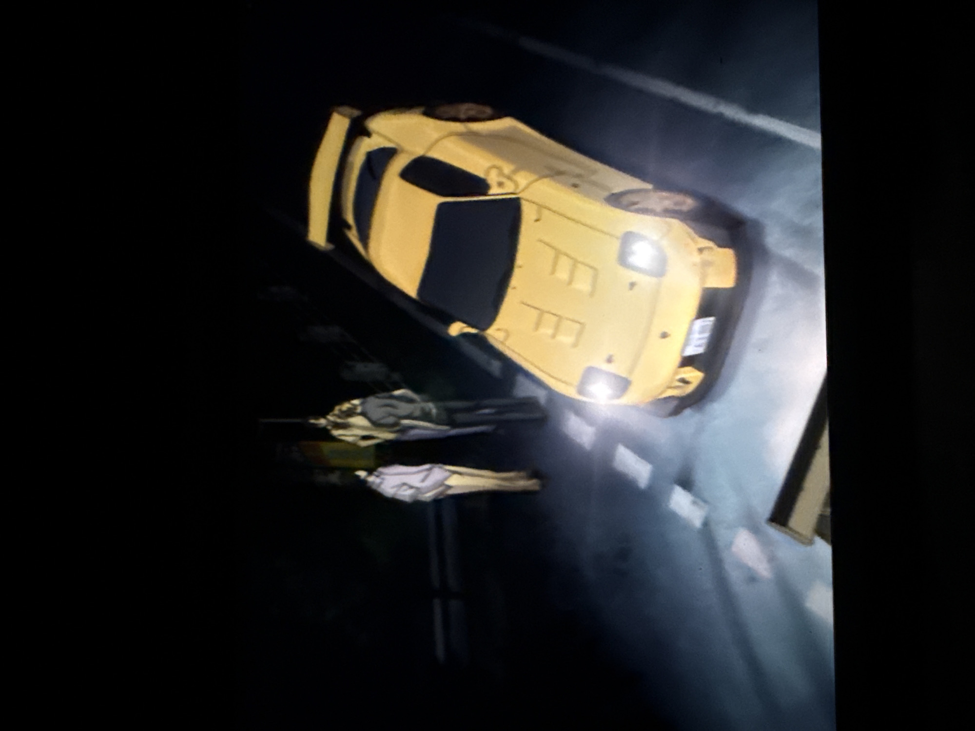 A yellow Mazda RX-7 is parked on a dark street at night. Two people are lying on the ground near the car. The location is unknown.