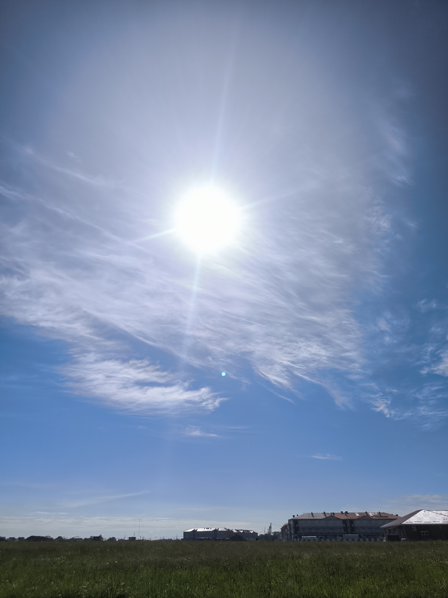 In the city of Laskino, Russia, on a clear, sunny day, a vast expanse of green grass dominates the foreground. In the mid-ground, a series of buildings are visible, some appearing to be under construction with scaffolding. The sky above is a vibrant blue, with wispy white clouds streaking across it. The sun, a bright white orb, is positioned high in the sky, emitting strong rays.