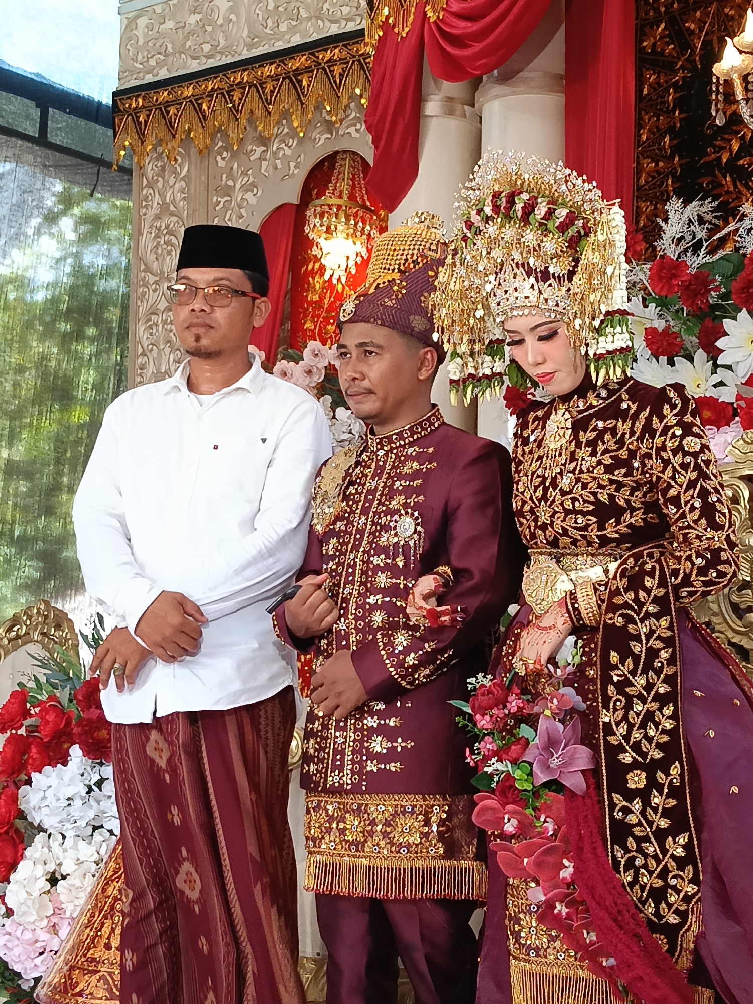 Three individuals are present at what appears to be a wedding ceremony in Geulumpangsulu Timu, Indonesia. On the left, a man wearing a white long-sleeved shirt, a black peci (cap), and glasses stands with his hands clasped in front of him. He is also wearing a patterned maroon sarong. In the center, another man is dressed in a maroon traditional suit adorned with gold embroidery and a matching cap. He stands with his left arm extended, hand clasped with the woman to his right. The woman on the right is adorned in a highly ornate maroon traditional wedding outfit, including a large, elaborate gold and red headdress. Her outfit also features extensive gold embroidery. She is wearing gold jewelry and her hands are decorated with henna. Both the central man and the woman are standing close together, suggesting they are the couple being celebrated. They are positioned in front of a backdrop decorated with red curtains, gold trim, and floral arrangements of red and white flowers. The overall scene suggests a formal cultural event.