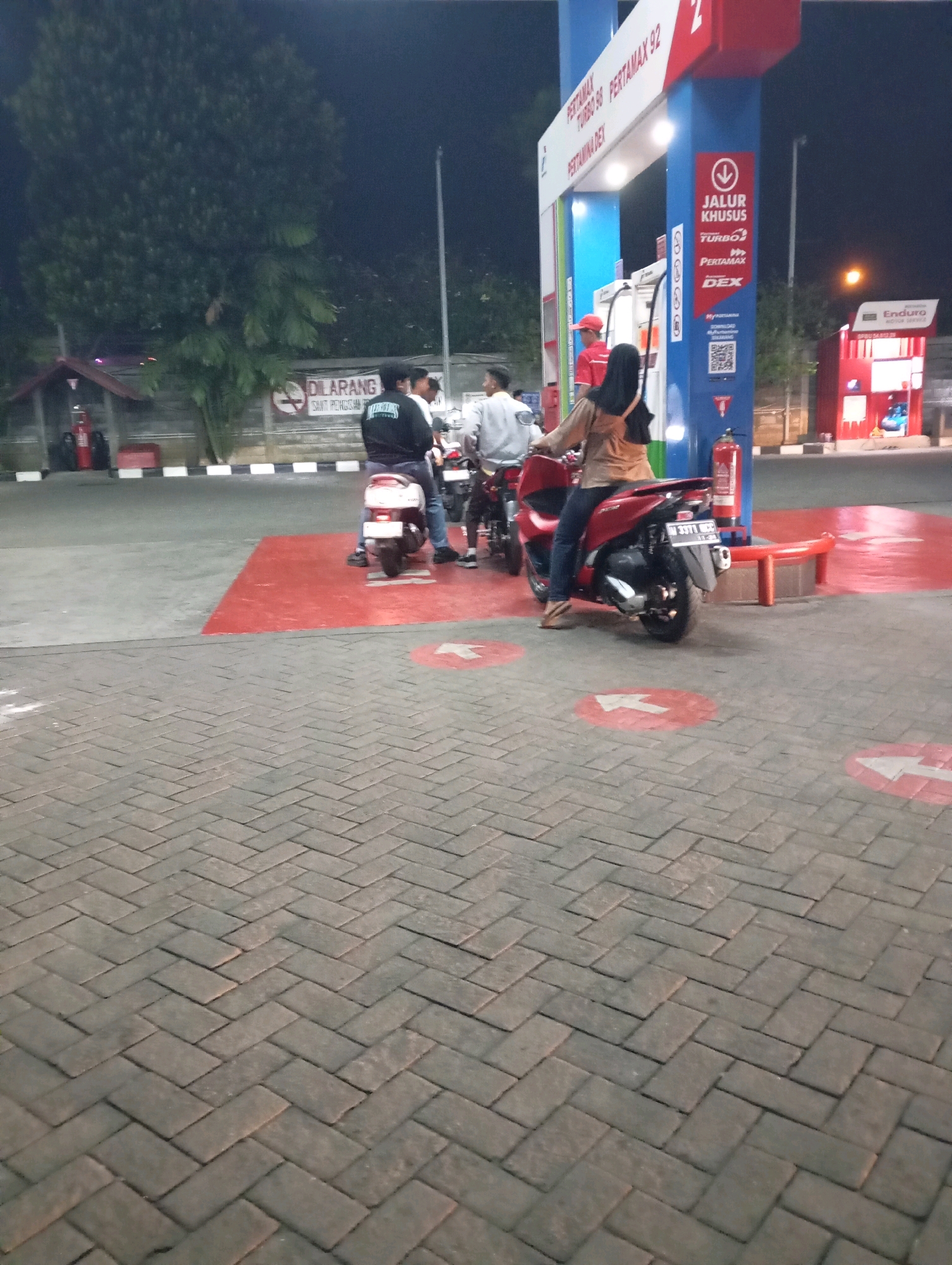 The image captures a nighttime scene at a Pertamina gas station in Ketegan, Indonesia. The focal point is a red scooter with a female rider wearing a hijab and brown jacket, positioned in front of a gas pump. To the left, a white scooter is visible with two individuals, one in a black jacket. A gas station attendant in a red shirt and cap stands near the pump. The ground is paved with interlocking bricks, and red circular markings with white arrows indicate traffic flow. A sign reading "DILARANG" (Forbidden) with a cigarette icon is present on a wall in the background, along with a fire extinguisher mounted on the gas pump structure. The lighting suggests it is after dark, with the gas station illuminated by artificial lights.