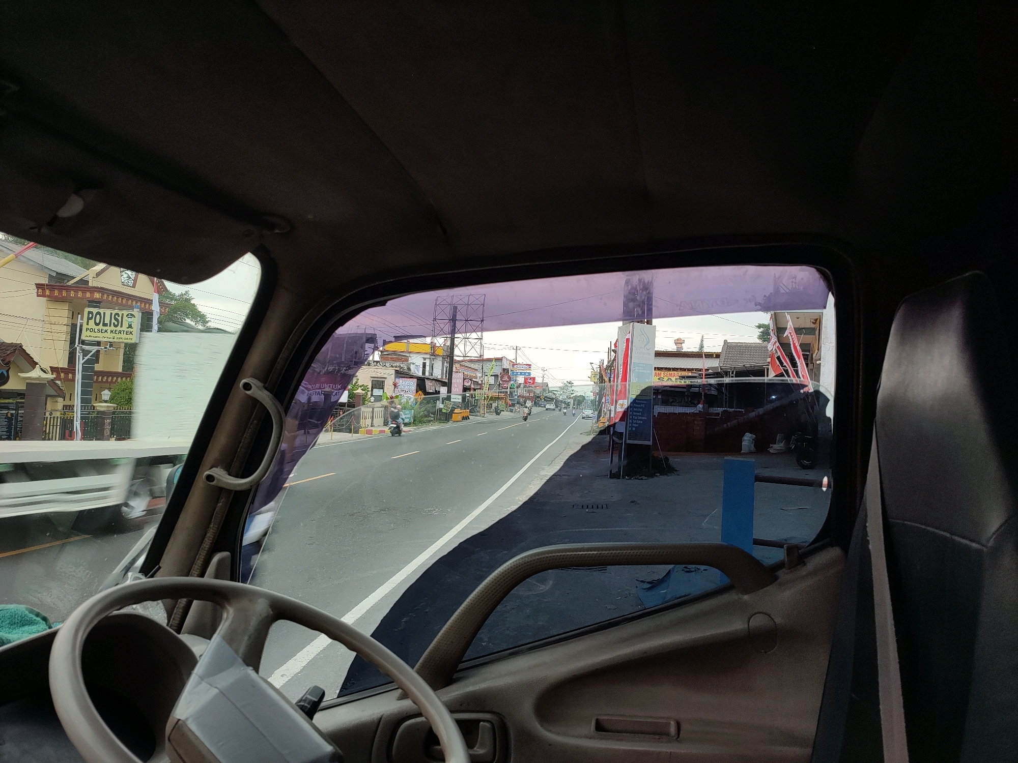 The image is taken from the interior of a vehicle, looking forward through the windshield. A steering wheel is visible in the lower left corner. The view outside shows a street in Karangluhur, Indonesia. Buildings line both sides of the road, with a police station identified by a sign reading "POLISI POLSEK KERTEK" on the left. Vehicles, including motorcycles and cars, are present on the road. The sky is overcast. The overall impression is that of a street scene in a small Indonesian town during daylight.