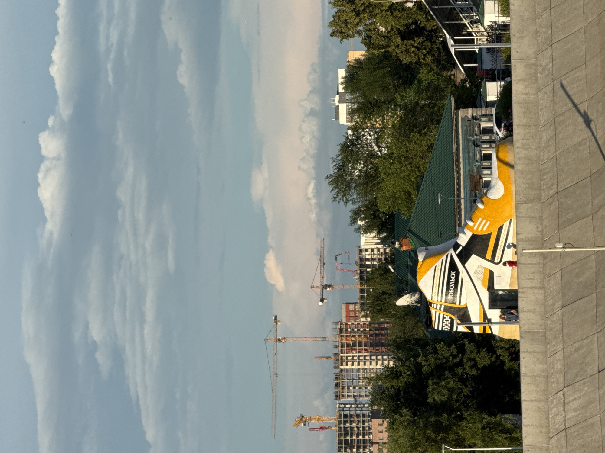 The image depicts a section of the city of Tyumen, Russia, with a prominent building shaped like a giant yellow and white sneaker as the central subject. The sneaker-shaped building is situated behind a concrete retaining wall and surrounded by trees. The word "ТОБОЛЬСК" is visible on the side of the sneaker. Two individuals, one in a blue top and another in a red top, are standing near the entrance of the building.
In the background, construction cranes and unfinished buildings indicate ongoing urban development. The sky is filled with dramatic clouds, suggesting either an approaching storm or the aftermath of one, with patches of sunlight illuminating parts of the scene. The overall impression is one of a blend of artistic architectural design and industrial progress within a city setting.