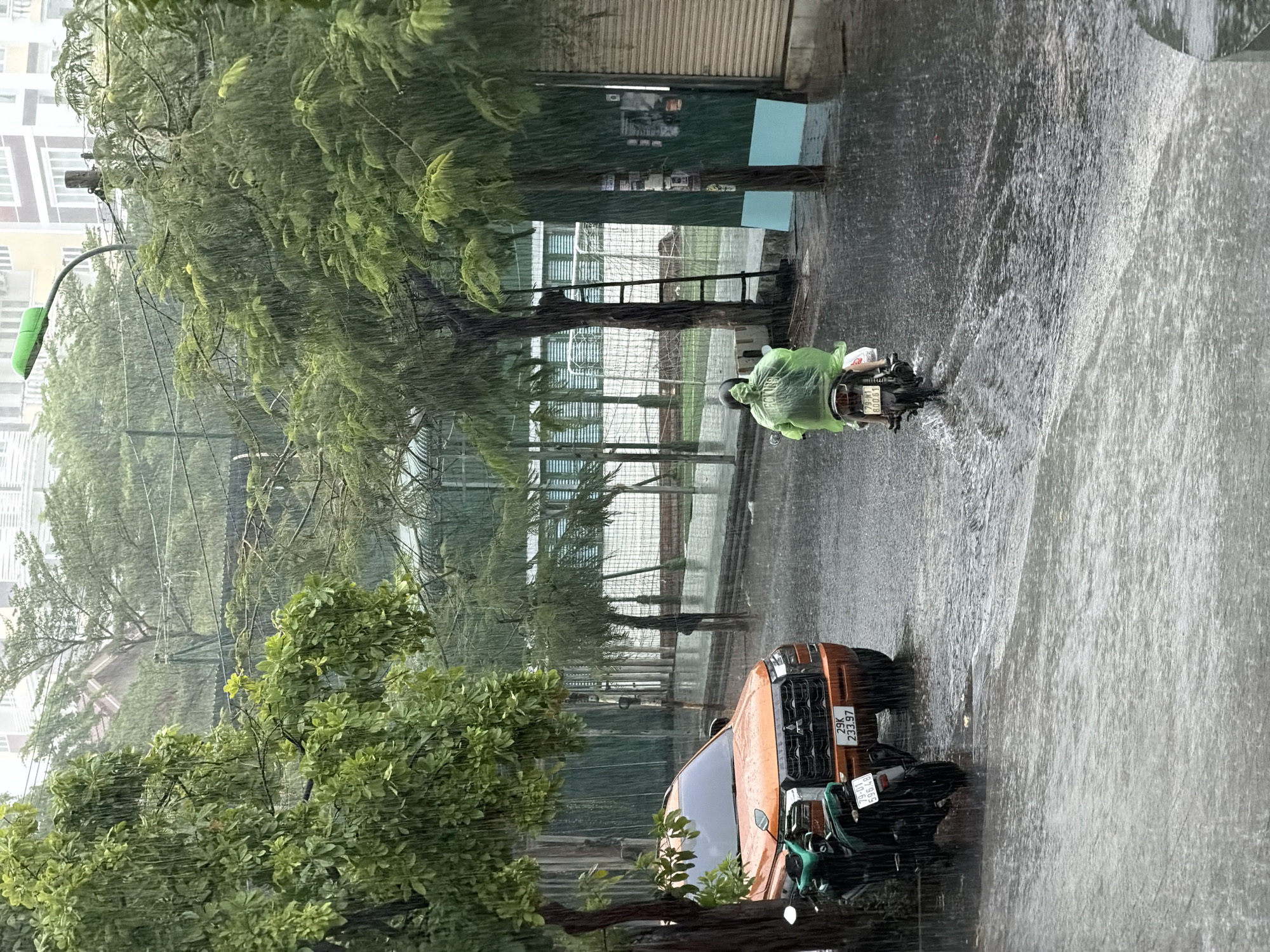 The image captures a street scene in Bac Nha Trang Ward, Vietnam, during heavy rain. A person wearing a green poncho is riding a motorcycle on the flooded street. An orange car and another motorcycle are parked on the side of the road. Trees line the street, and a building is visible in the background. The rain is falling heavily, obscuring some details of the scene.
